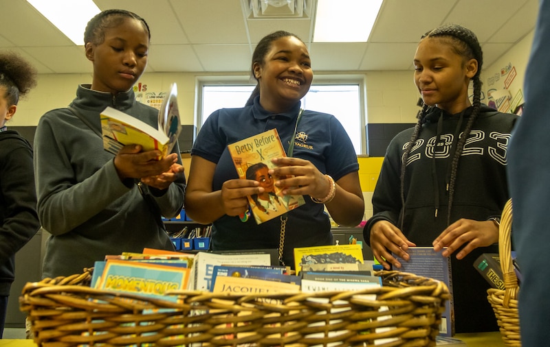 Three students are standing in front of a large basket of books. Two of the students are holding books.