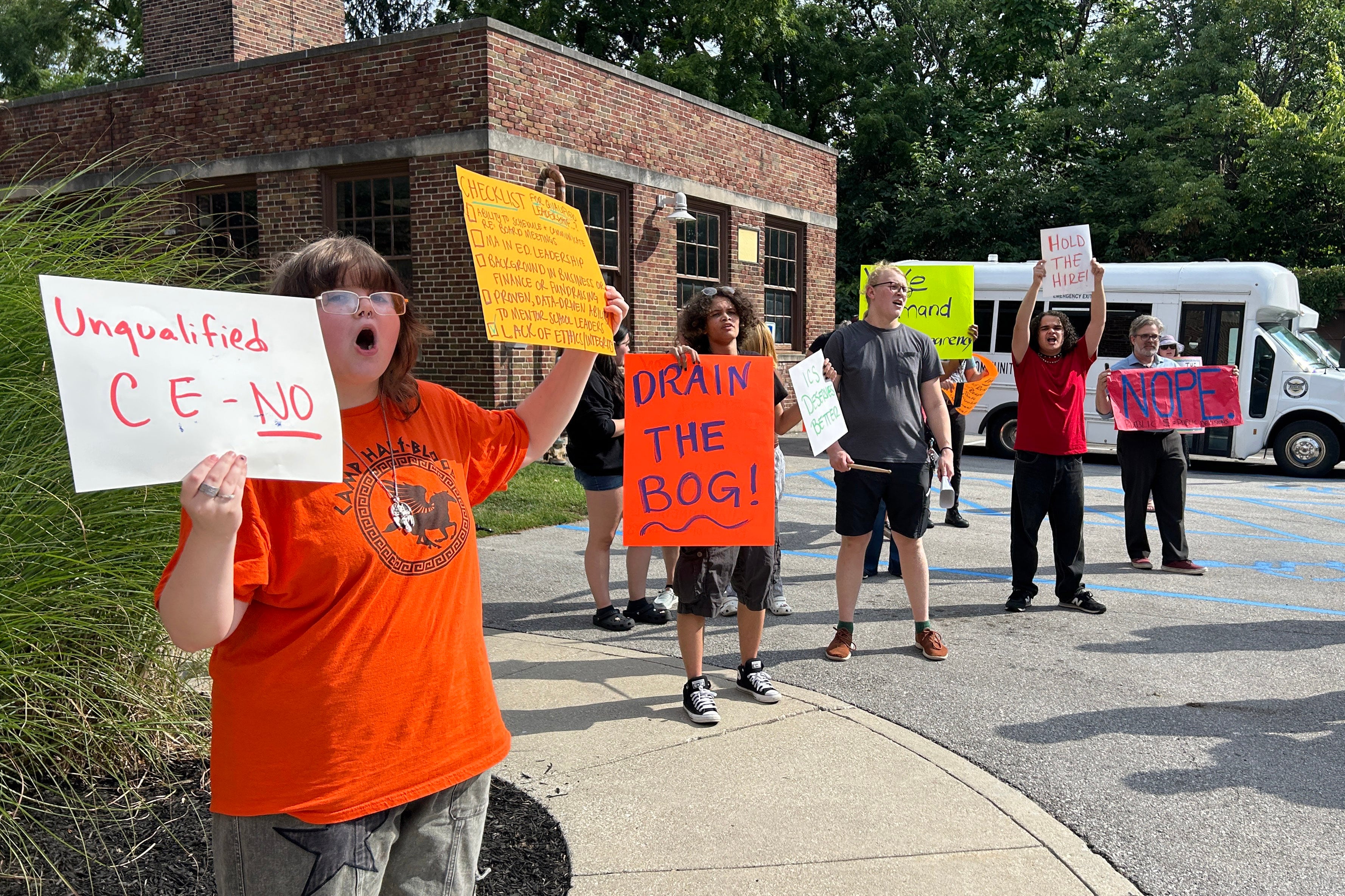 A photograph of a large group of protestors, some holding large neon paper signs stand outside.