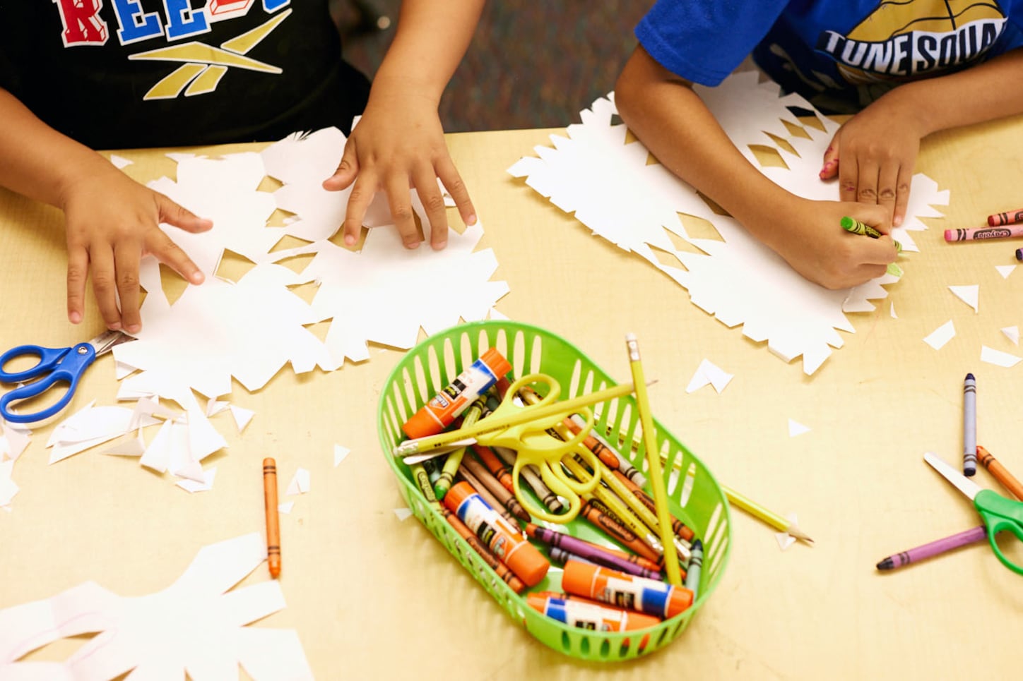 An up-close photo of two young students making white paper snowflakes with scissors and crayons.