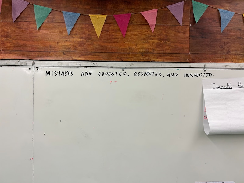 A whiteboard in a classroom that says "Mistakes are expected, respected, and inspected." There is a colorful triangle banner hanging above it.