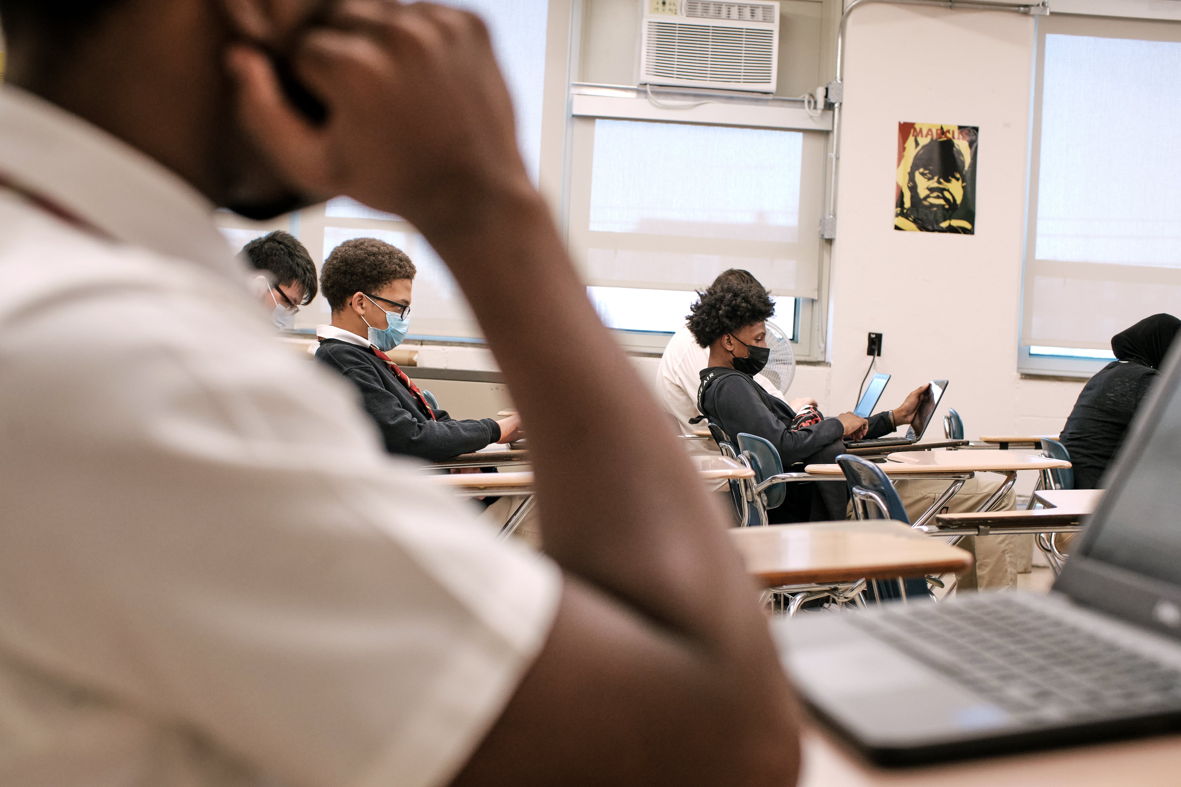 A couple of high school students wearing masks and sitting at their desks in a classroom all have a laptop in front of them with windows on the back wall in the background.