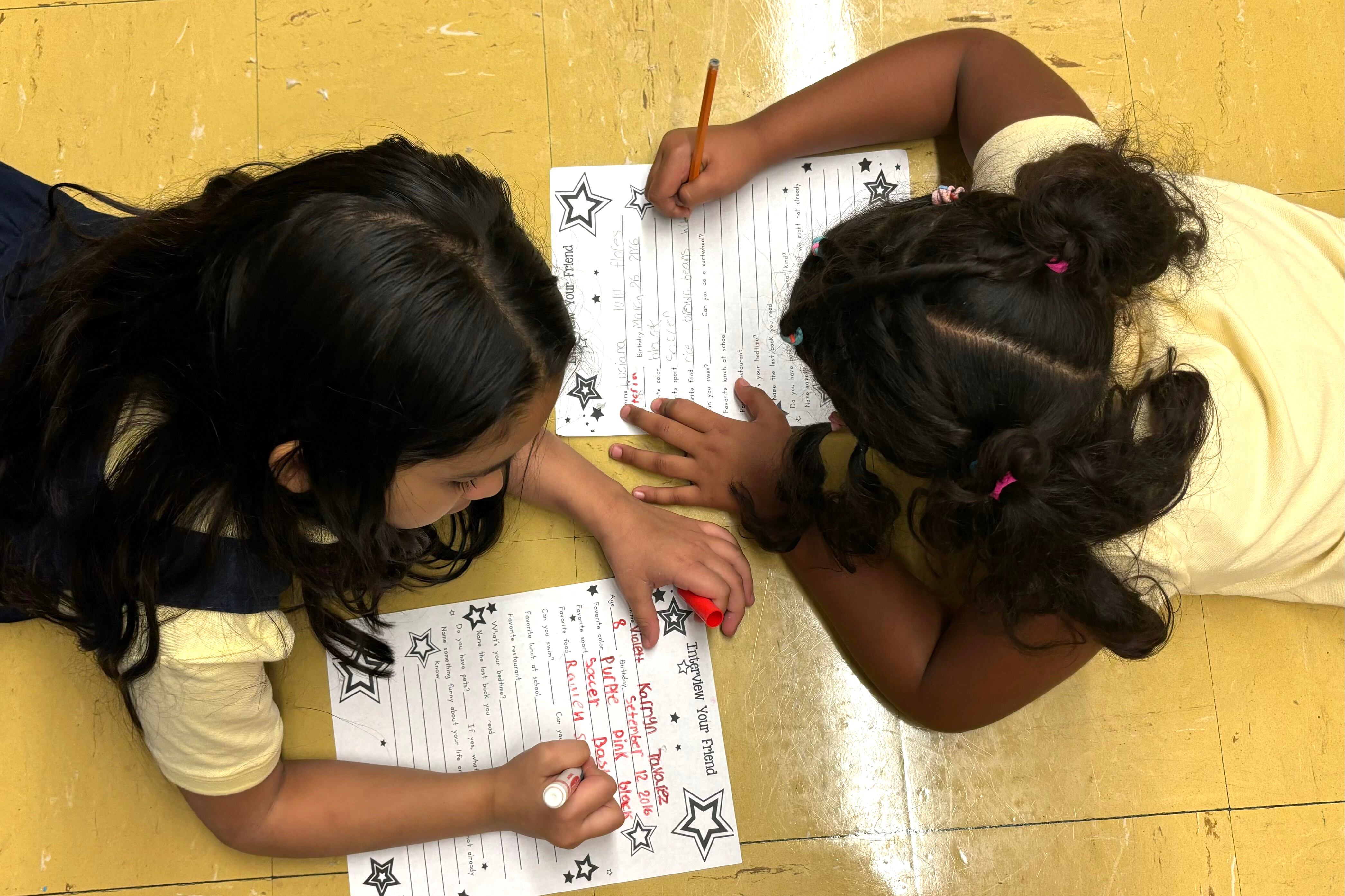 Two female students lie on the floor and work on papers.