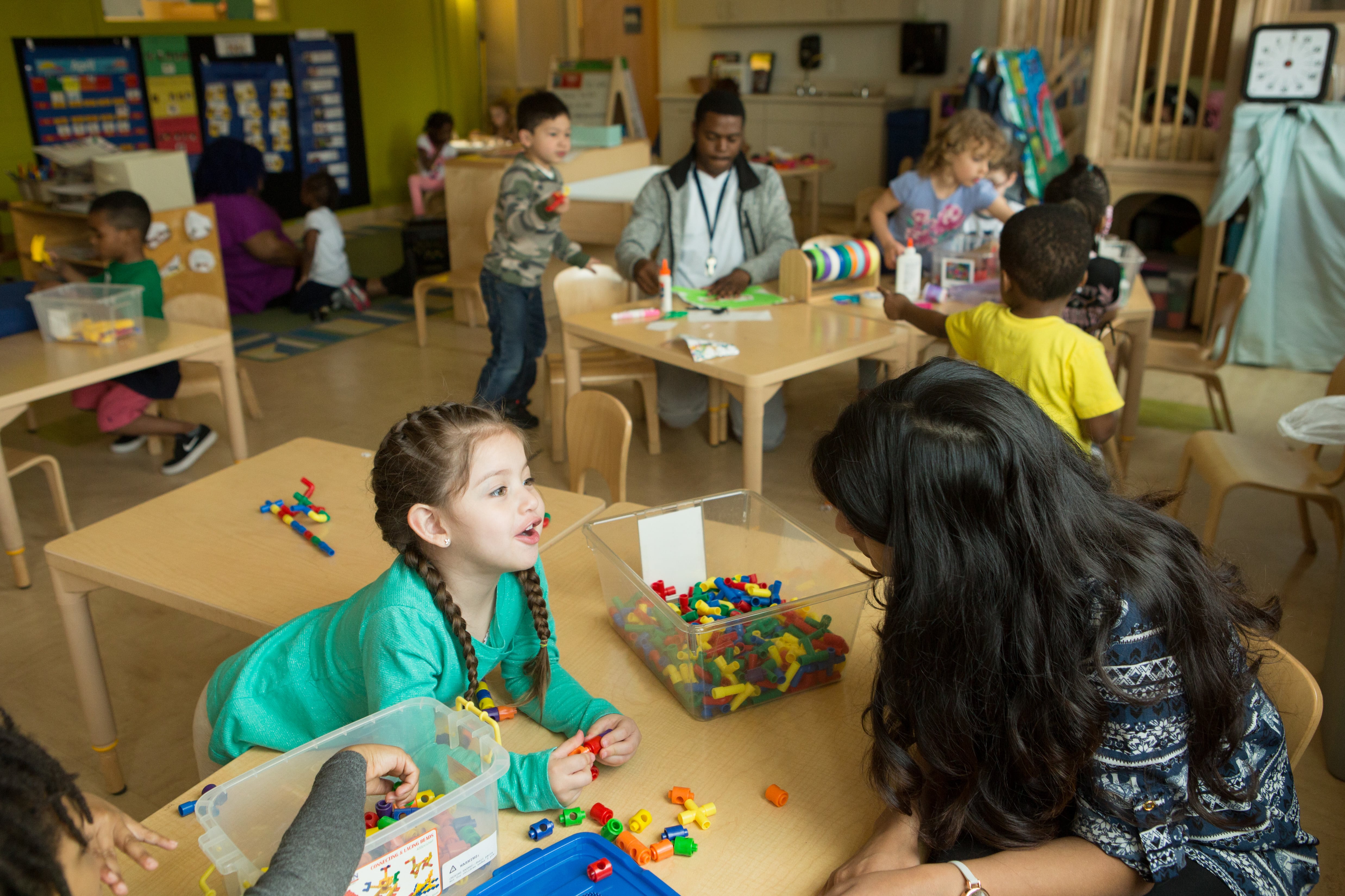 In a preschool class, a girl wearing a green shirt and pigtail braids leans over a desk, where there’s a bin full of building blocks, to talk to a woman who also leans in to listen.