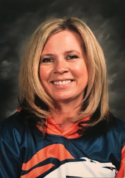 A woman with short, blonde hair wearing a Broncos football shirt smiles at the camera with a grey background.