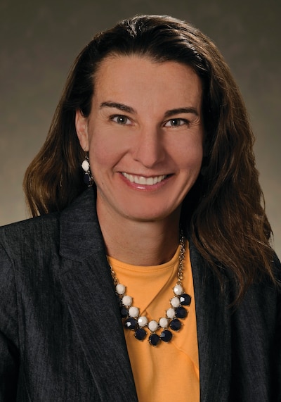 A woman with long dark hair. She is wearing an orange Tshirt, a black blazer, and a white and black bead necklace.