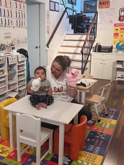 A woman holds up two hand puppets while supporting a small child in a daycare room with a colorful rug and toy and book shelves in the background.