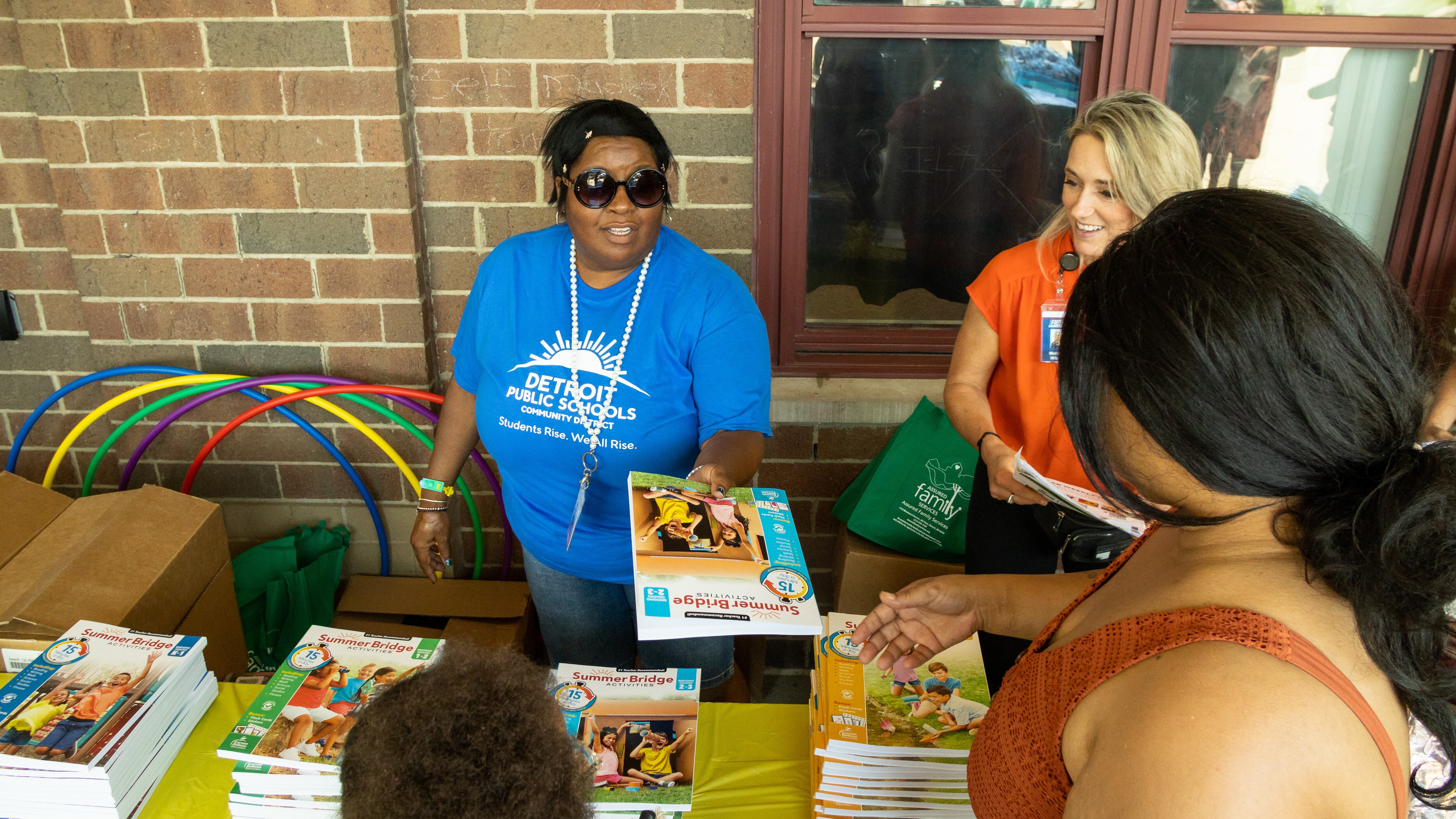 A woman standing behind a table hands curriculum materials to a parent who is accompanied by her child.