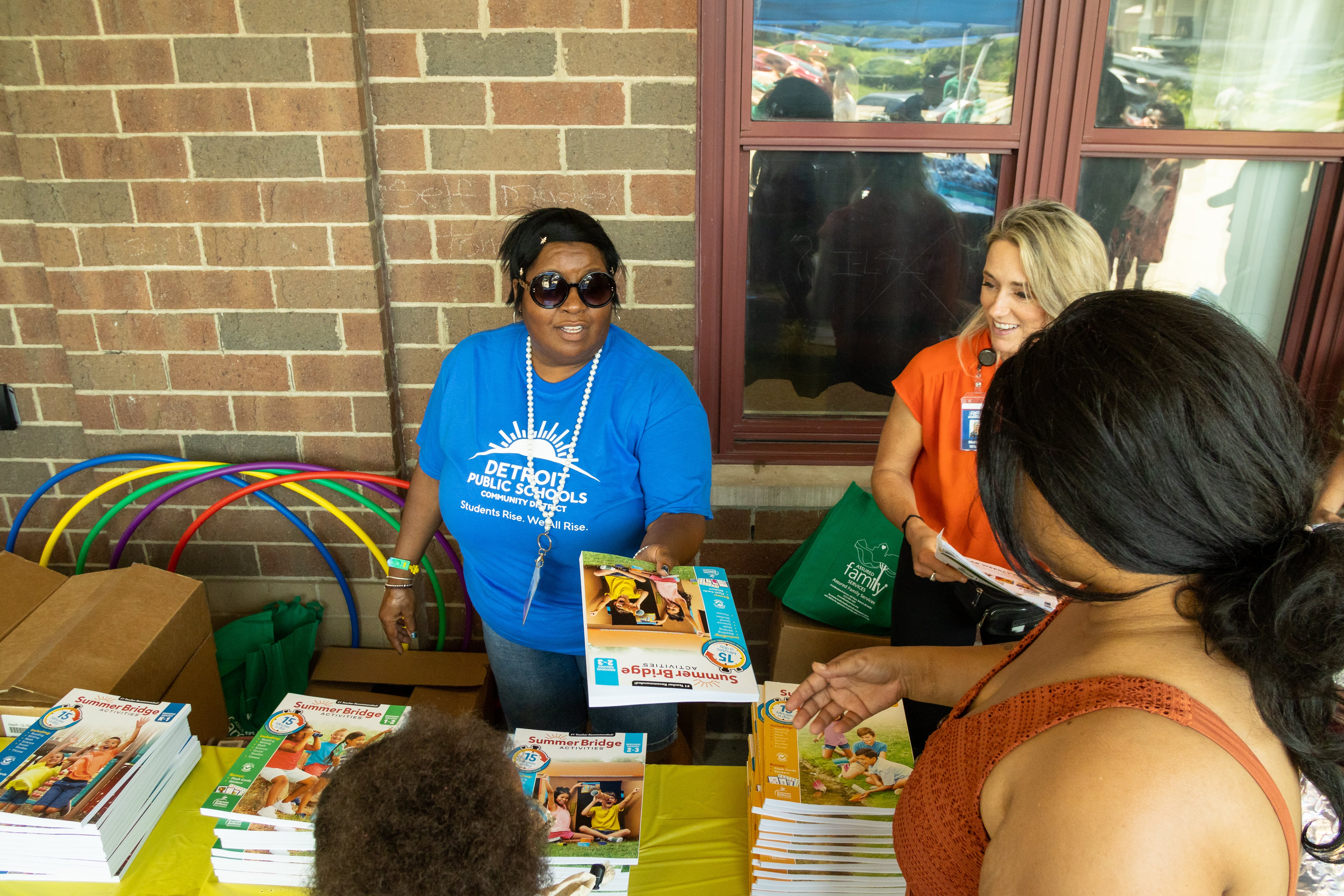 A woman standing behind a table hands curriculum materials to a parent who is accompanied by her child.