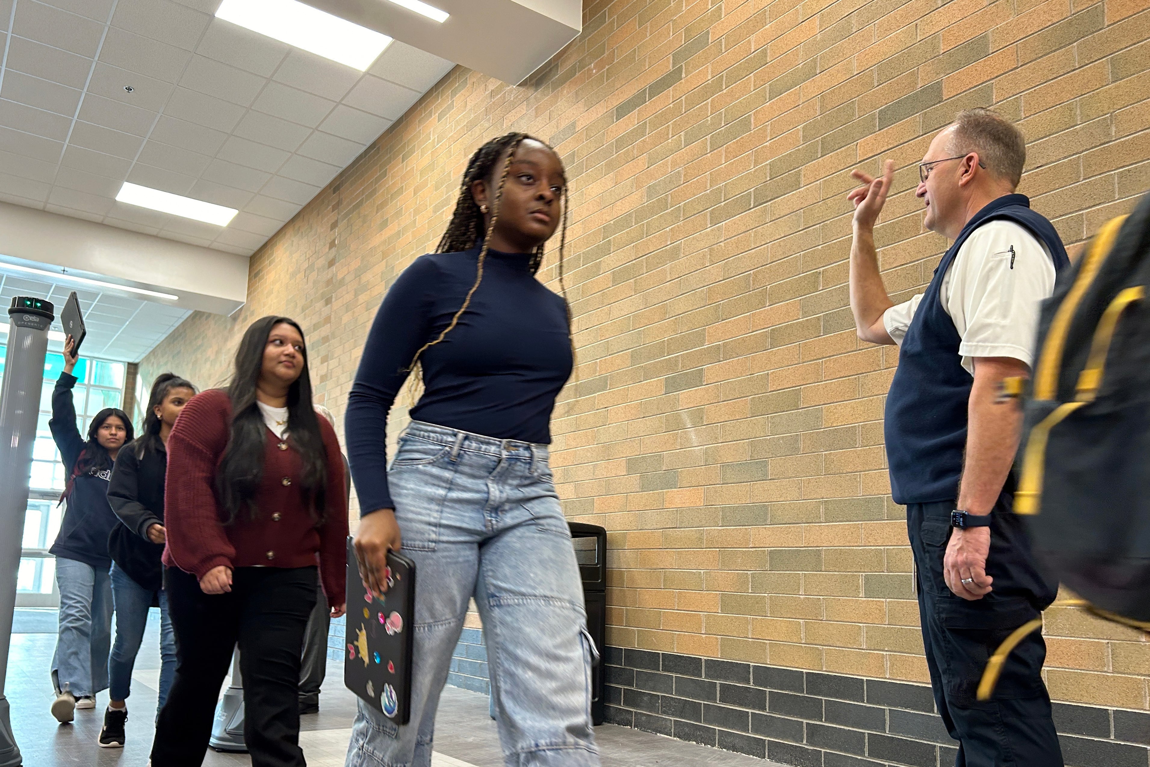 Students walk in a line past an adult security guard and metal detector with tan brick wall in the background.