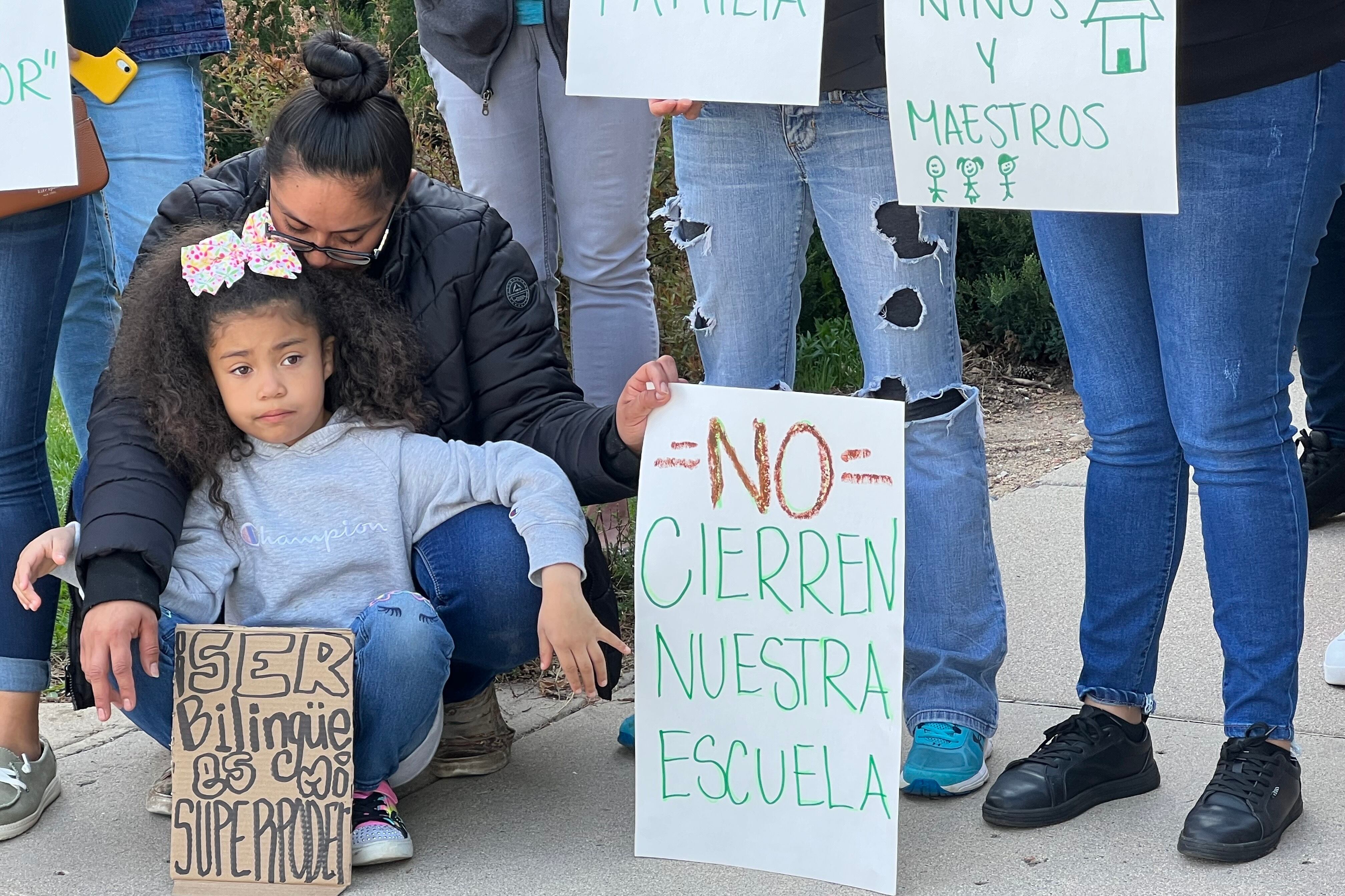 A little girl wearing jeans, a sweatshirt, and a hair bow holds a cardboard sign. In black marker, it says “Ser Bilingüe Es Mi Superpoder,” which means “Being Bilingual is my Superpower.”