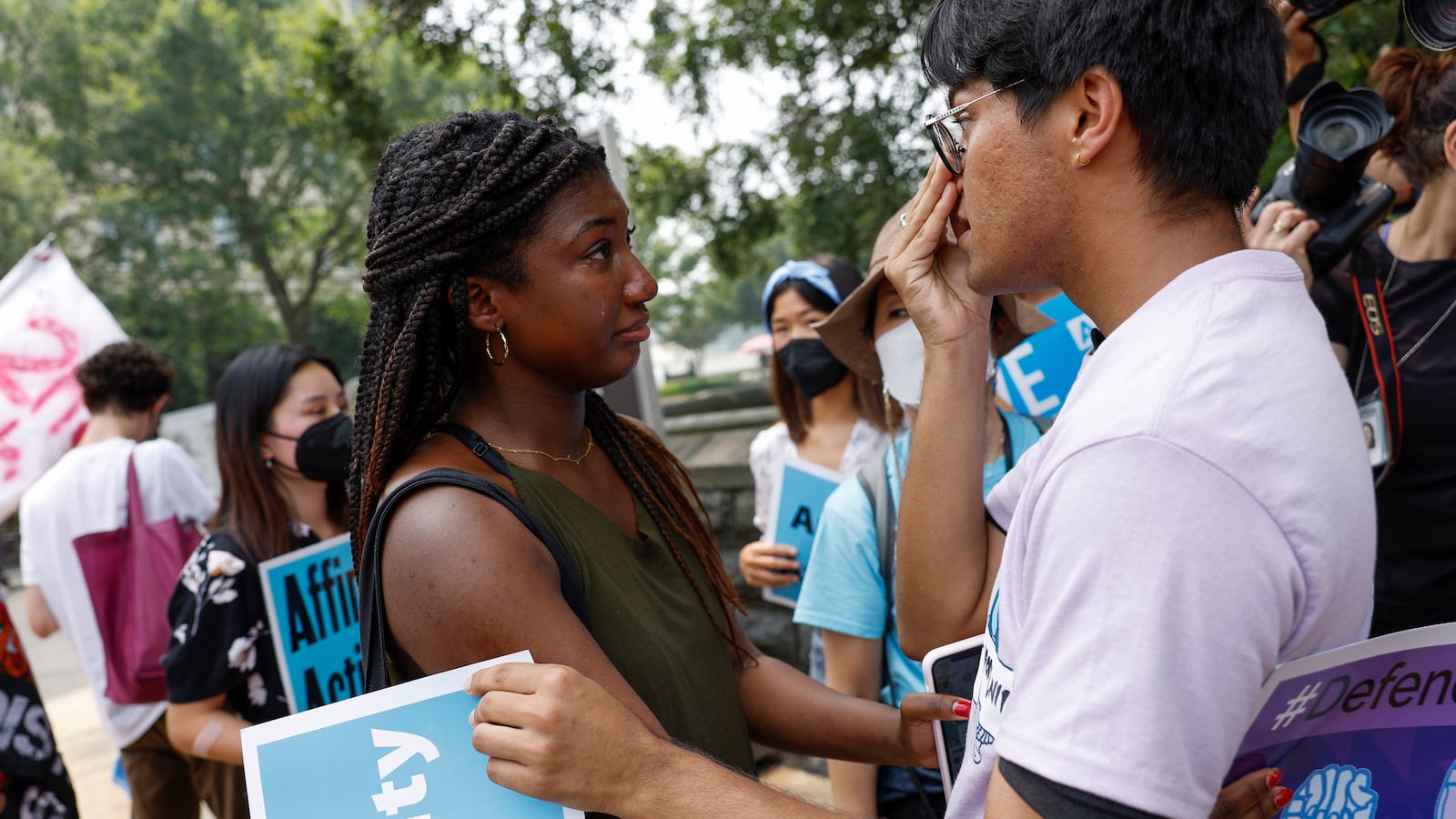 A young woman embraces a young man react with emotion to the news of the affirmative action ruling. They are carrying signs and embracing.