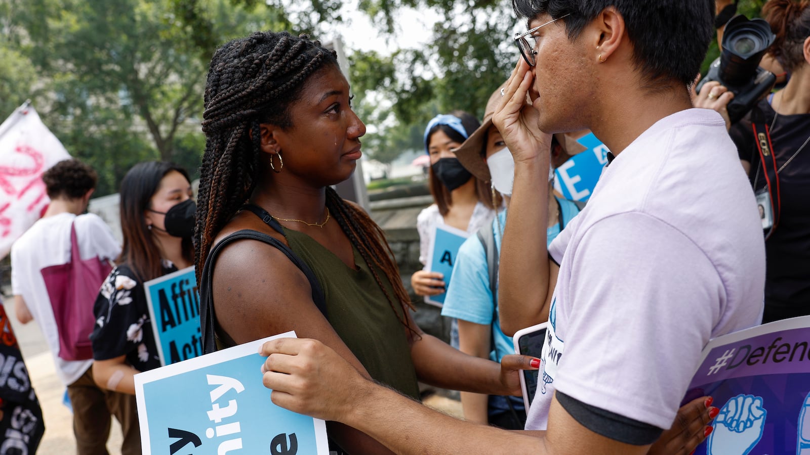 A young woman embraces a young man react with emotion to the news of the affirmative action ruling. They are carrying signs and embracing.