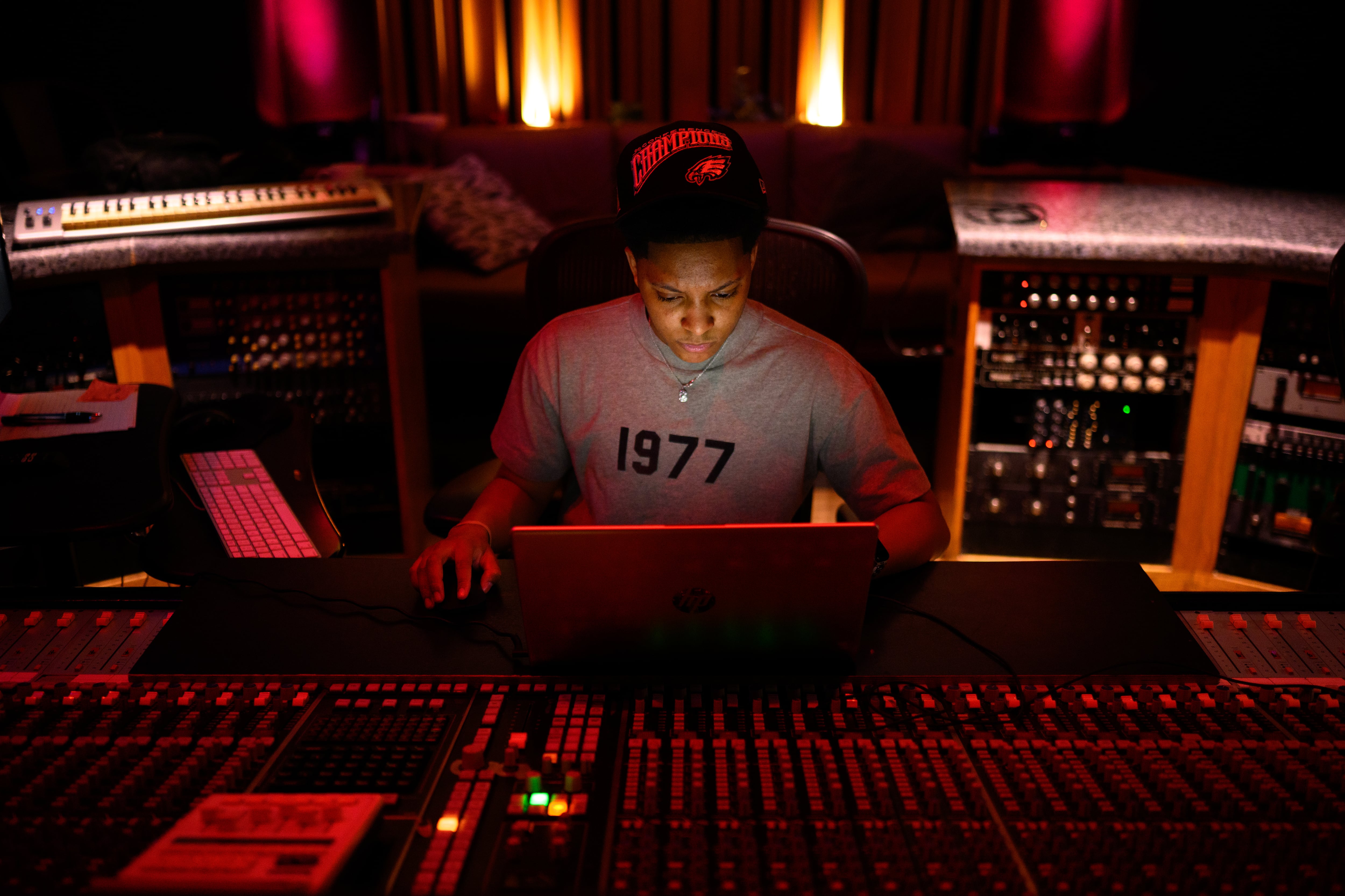 A high school Black student works inside of a recording studio in the dark with the colorful lights in the equipment.