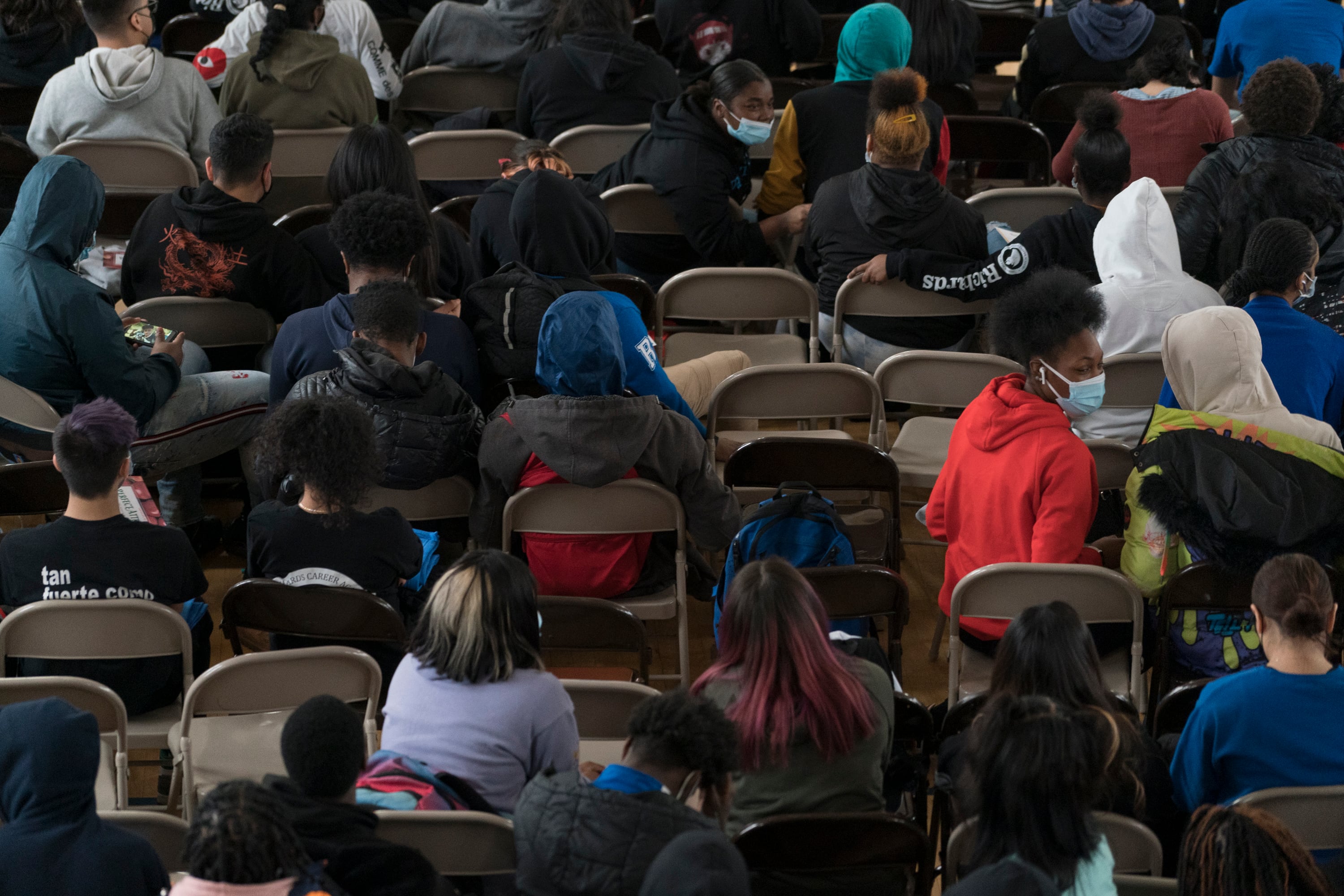 A large group of students sit in an auditorium together. Most of the students are sitting in beige chairs with blue face masks on turning to speak to each other.