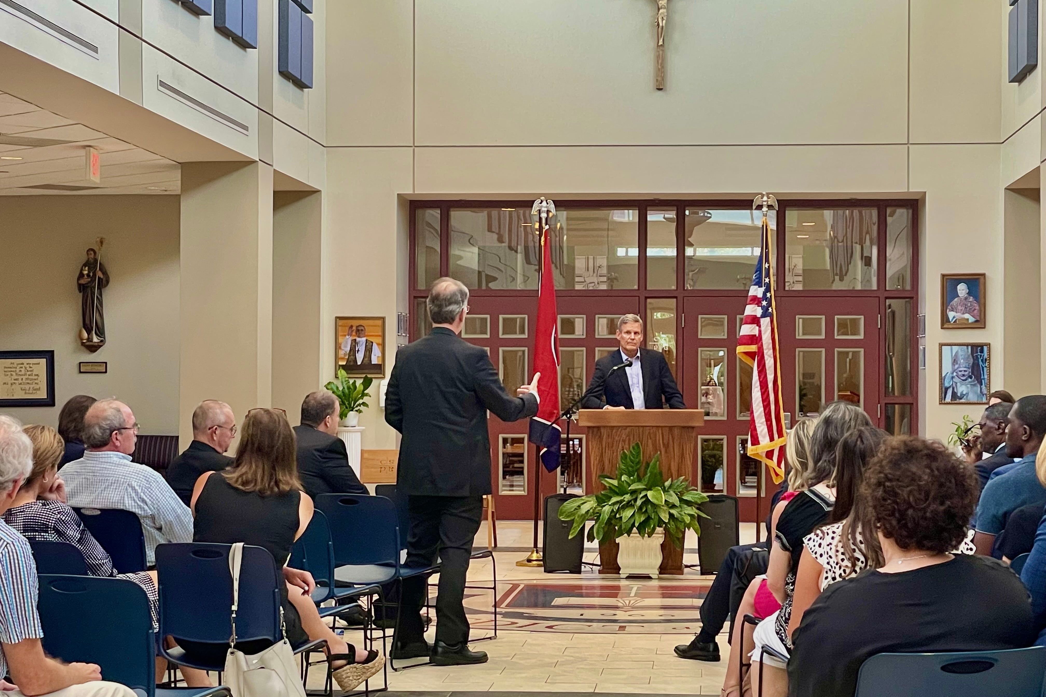 A man stands at a wooden podium, where a cross hangs above him, as he listens to another man ask a question, gesturing with his right arm.