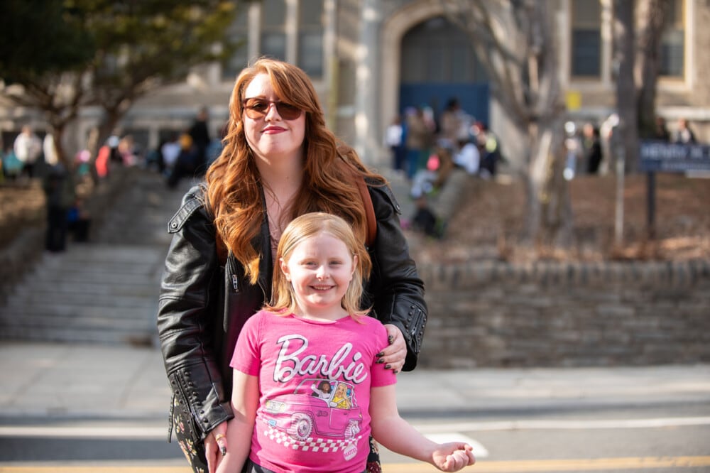 A portrait of a woman with sunglasses and a dark jacket and a girl with a pink Barbie t-shirt.