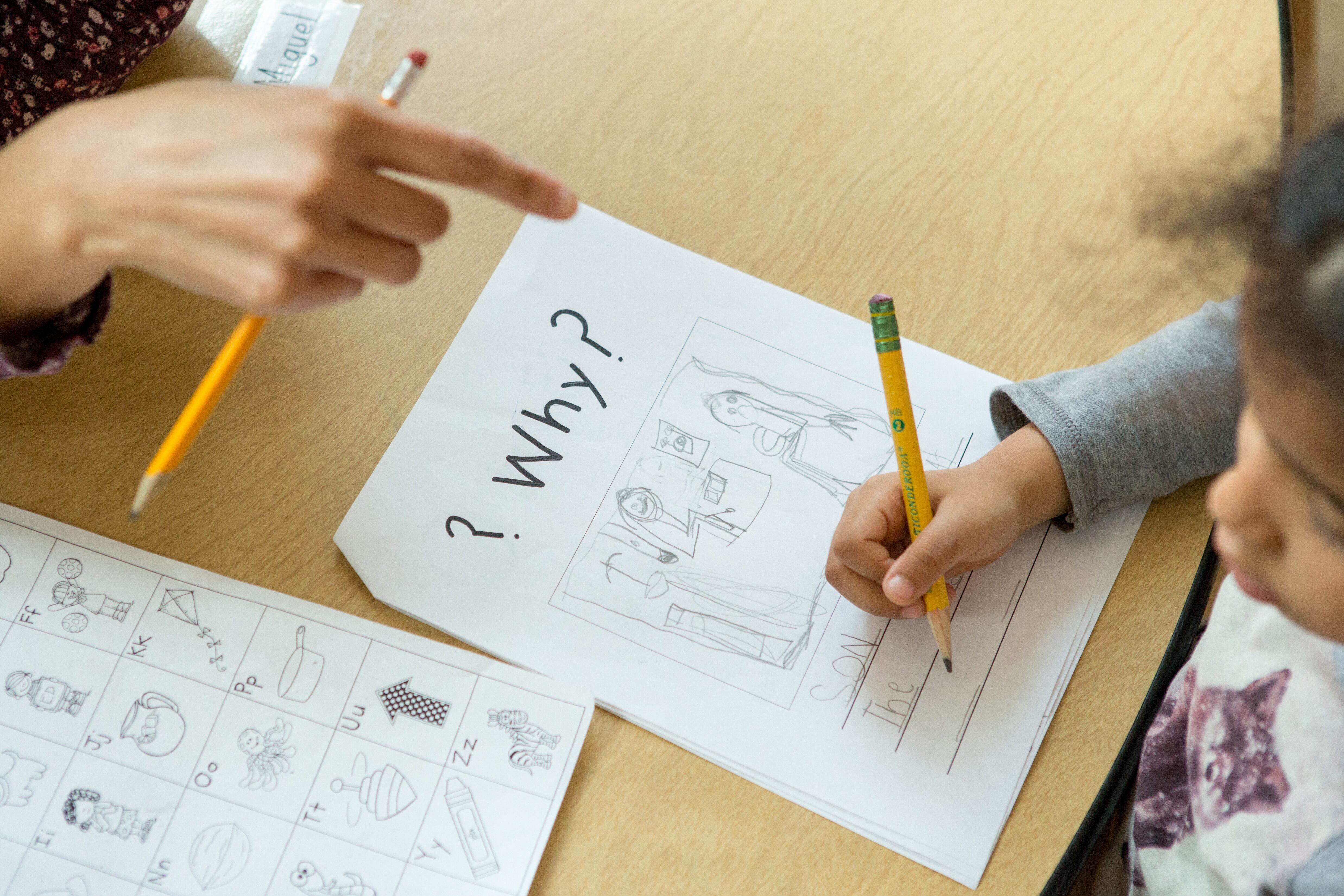 A teacher points to a kindergartener’s paper as she writes on it with a pencil. At the top of the worksheet, the word “Why?” is in bold black letters.