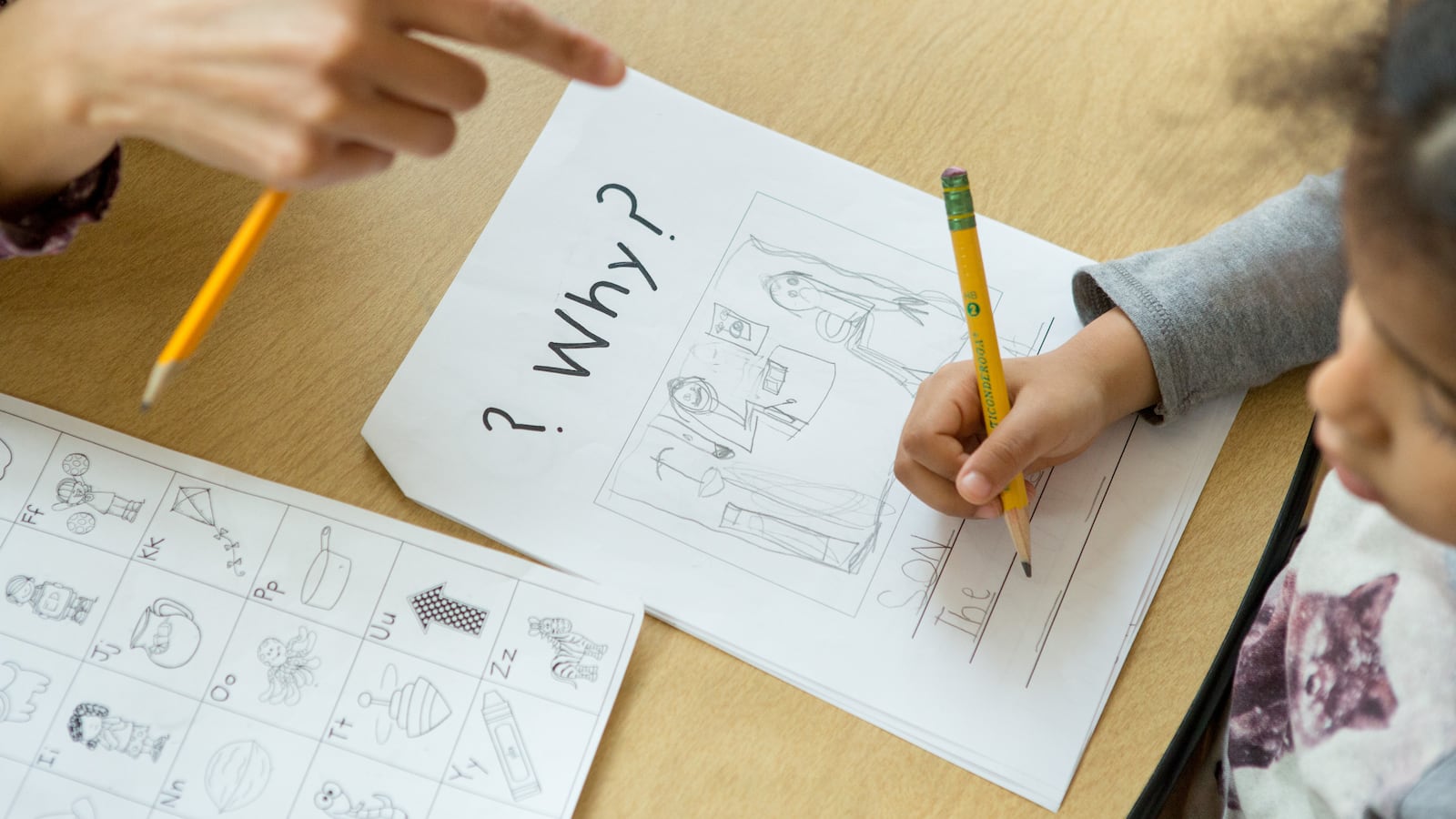 A teacher points to a kindergartener’s paper as she writes on it with a pencil. At the top of the worksheet, the word “Why?” is in bold black letters.