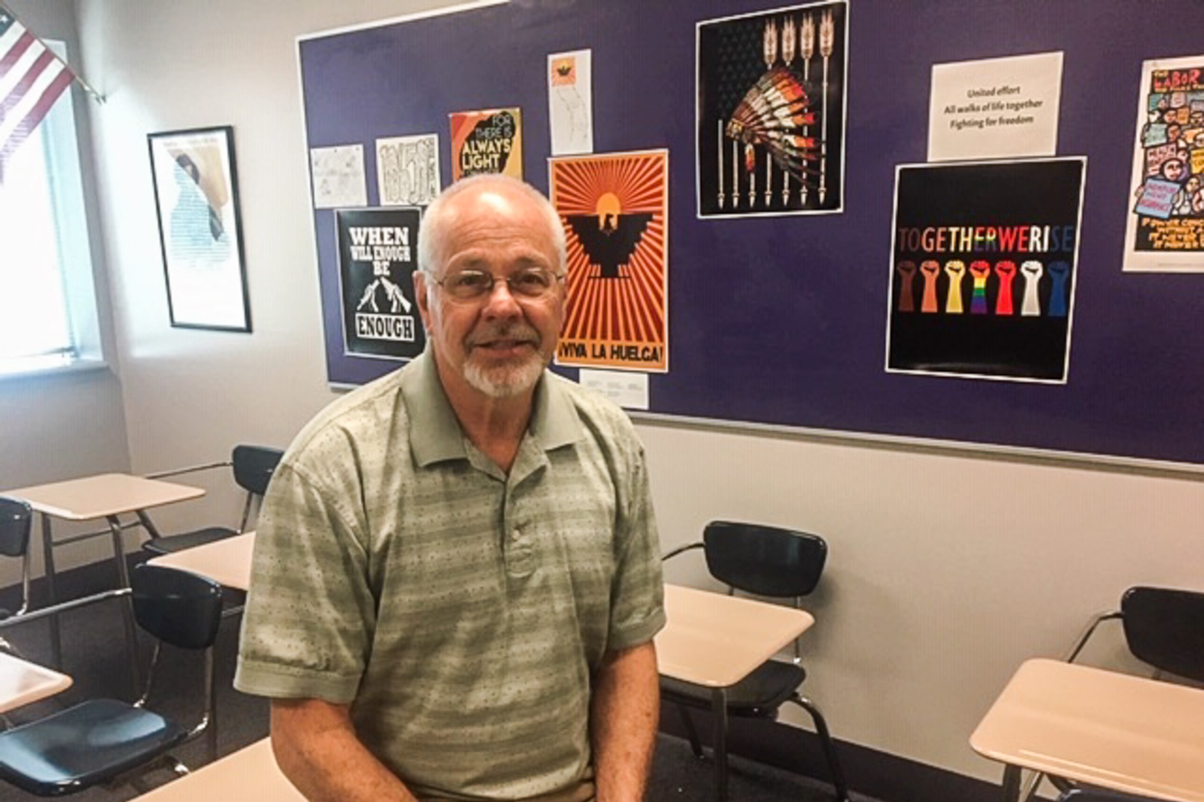 A high school history teacher sits on a desk in his classroom.