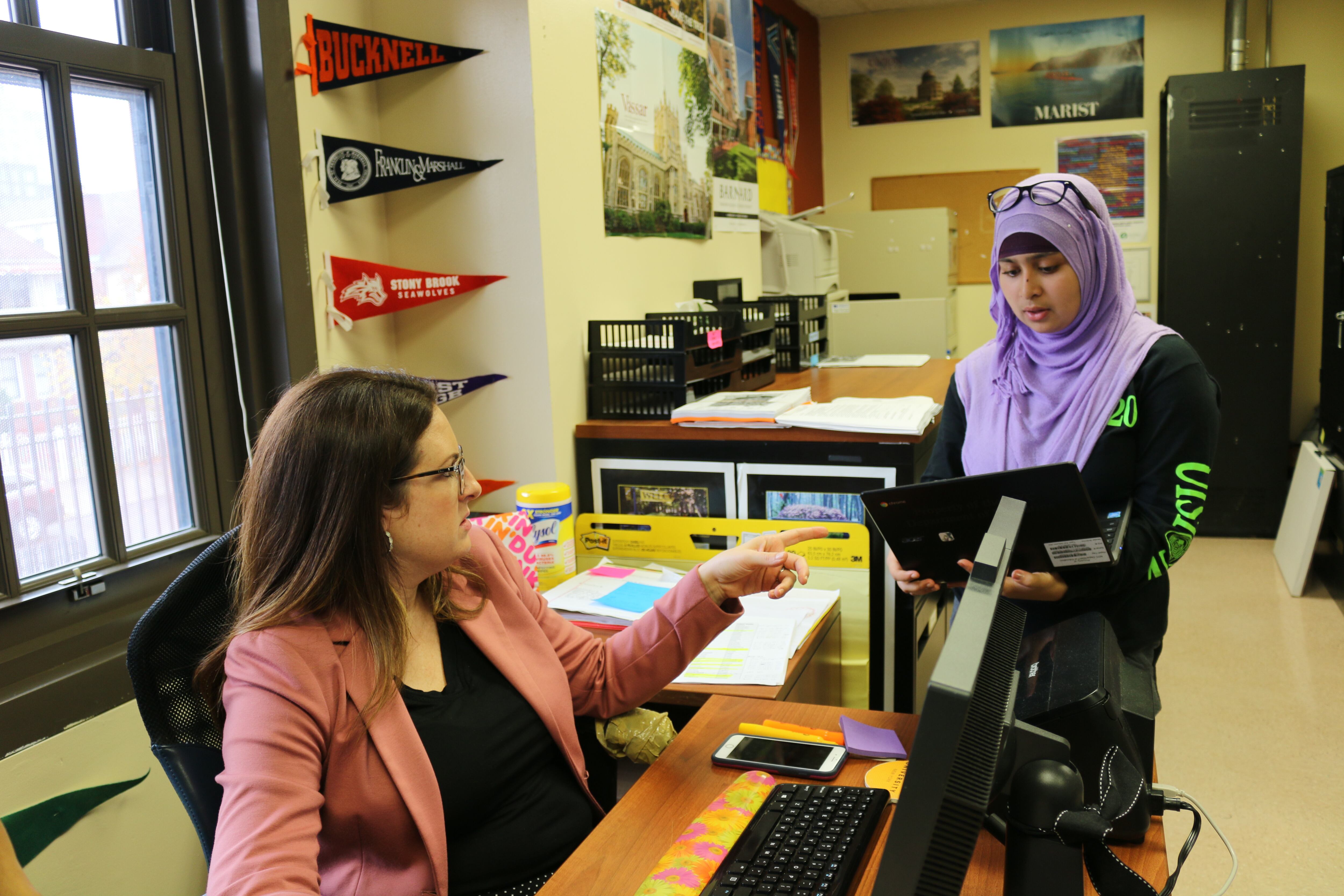 A woman wearing a light pink jacket sits at a desk while a student wearing a purple hijab stands next to the desk holding a laptop. The two are talking and there are college banners and posters on the wall in the background.
