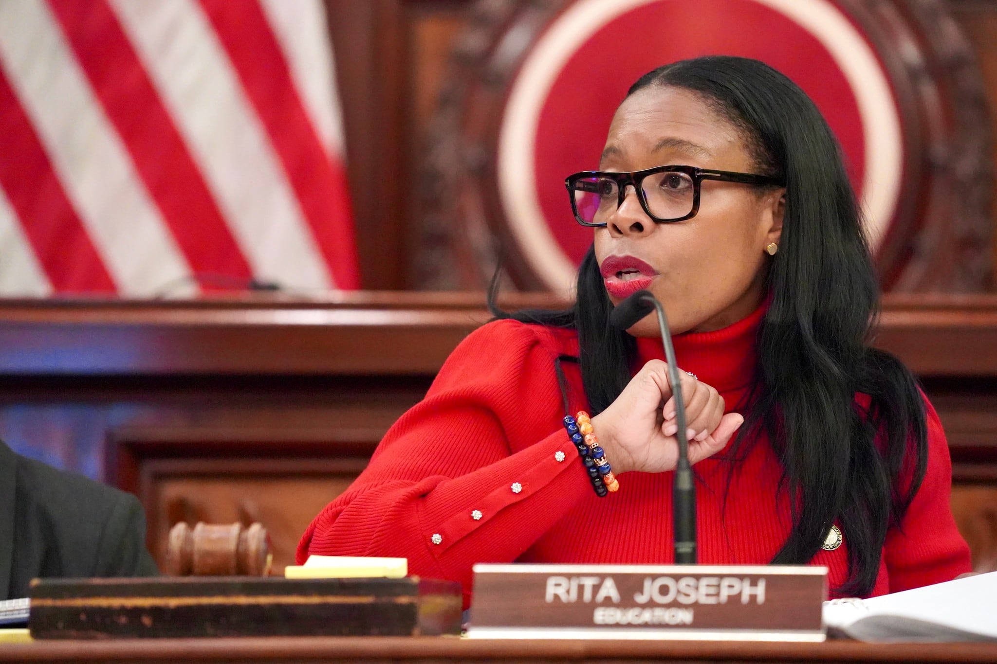 A woman wearing glasses and a red top speaks behind a nameplate.