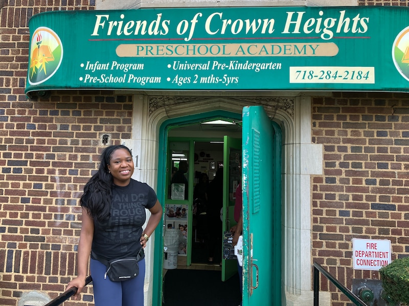 A Black woman wearing a black t-shirt and jeans smiles for a portrait outside of a school building with a green awning.