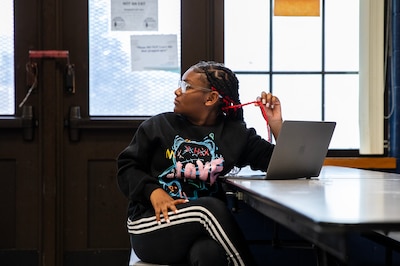 A young girl with long black hair looks over her right shoulder while twirling a pink braid while she sits at a table with a laptop in front of her.