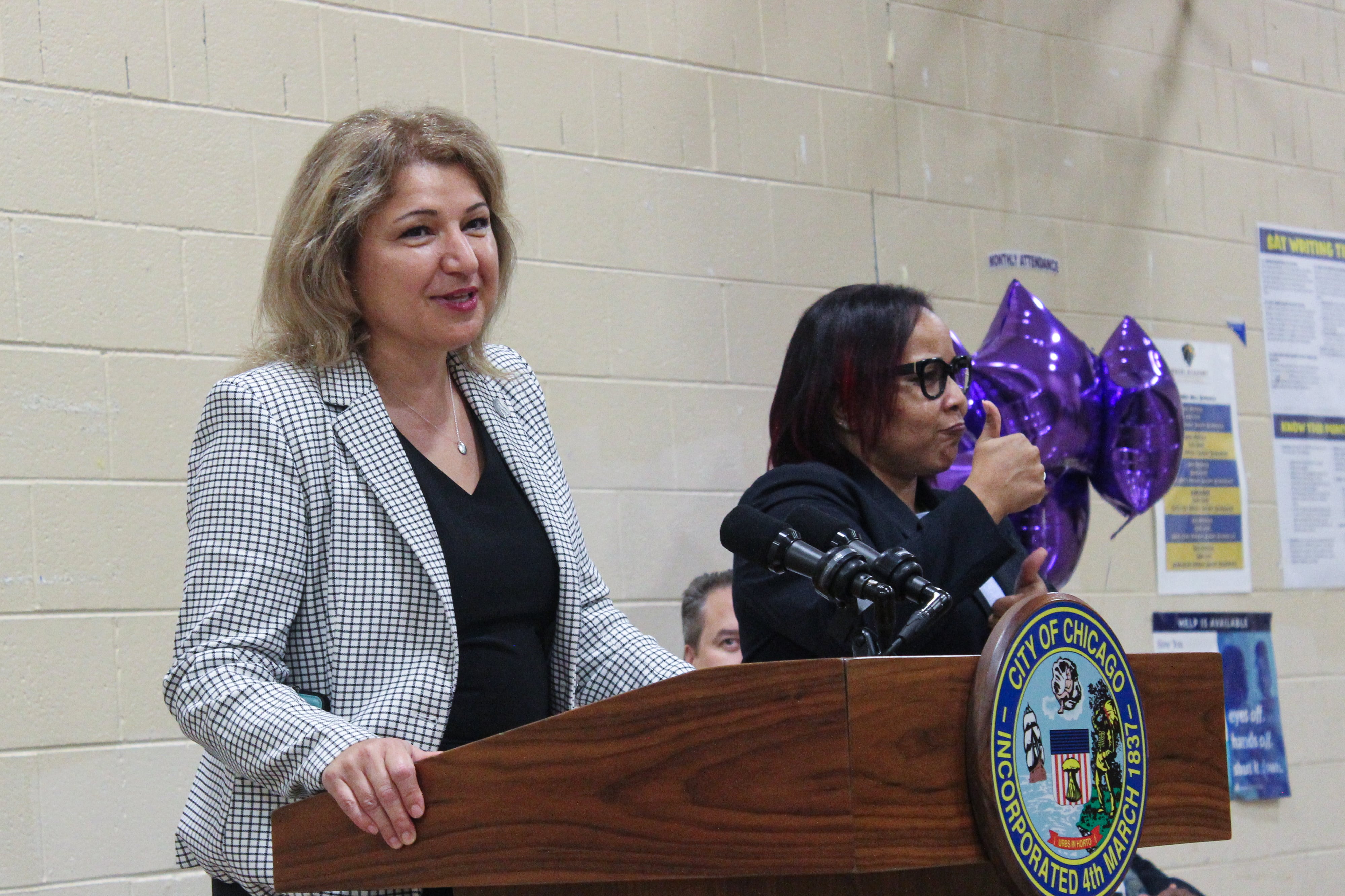 A woman in a sports coat stands at a lectern with another woman giving the thumbs-up nearby.