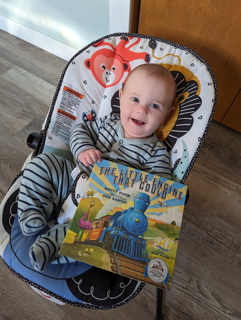A baby sits in a bouncy chair on wood floor with a giant picture book.