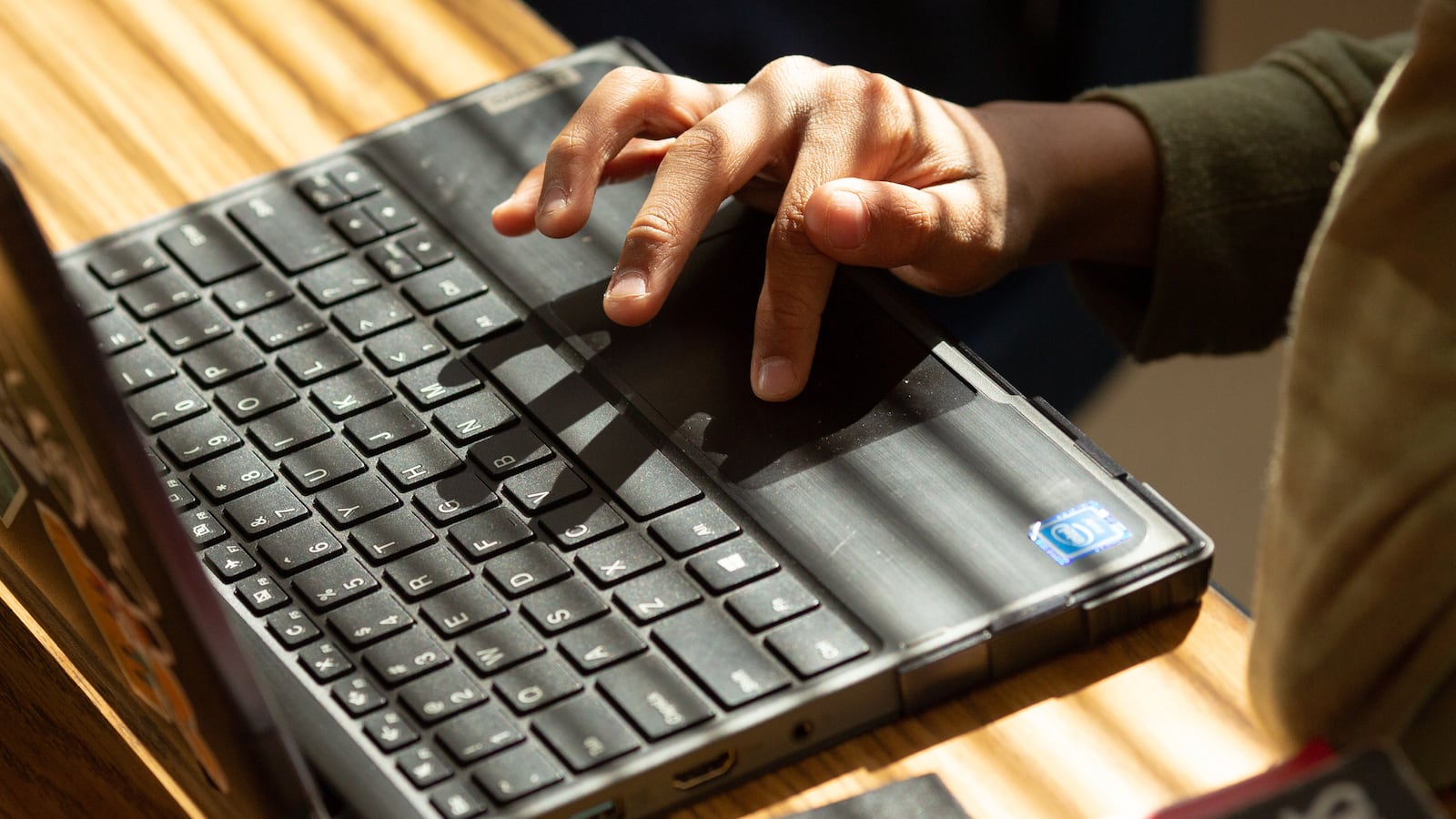 Someone’s hand types on a laptop as light shines through blinds onto the desk.
