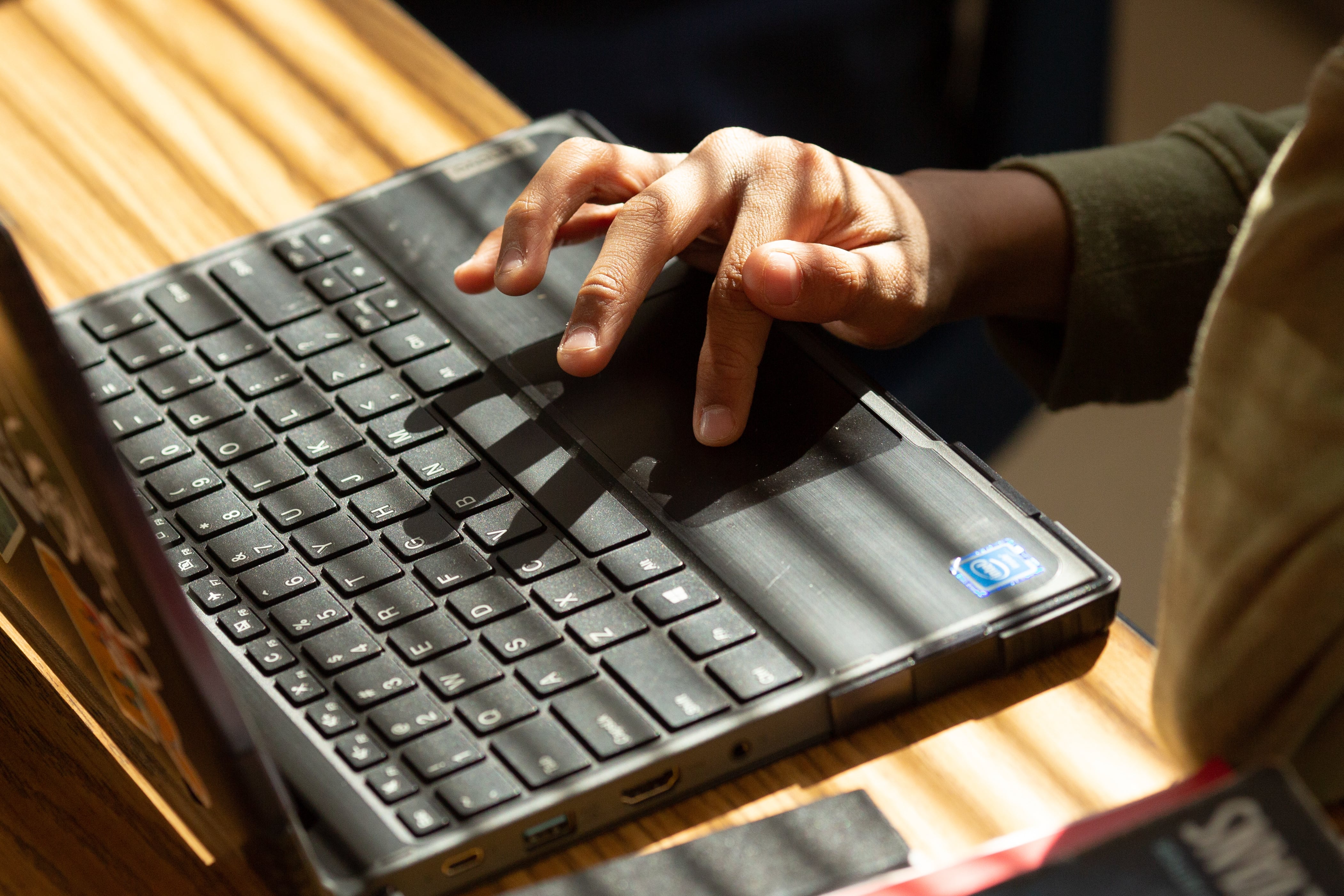 Someone’s hand types on a laptop as light shines through blinds onto the desk.