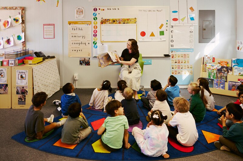 A teacher sits in a chair reading a book to a class full of young preschool students sitting on a colorful rug in a class.