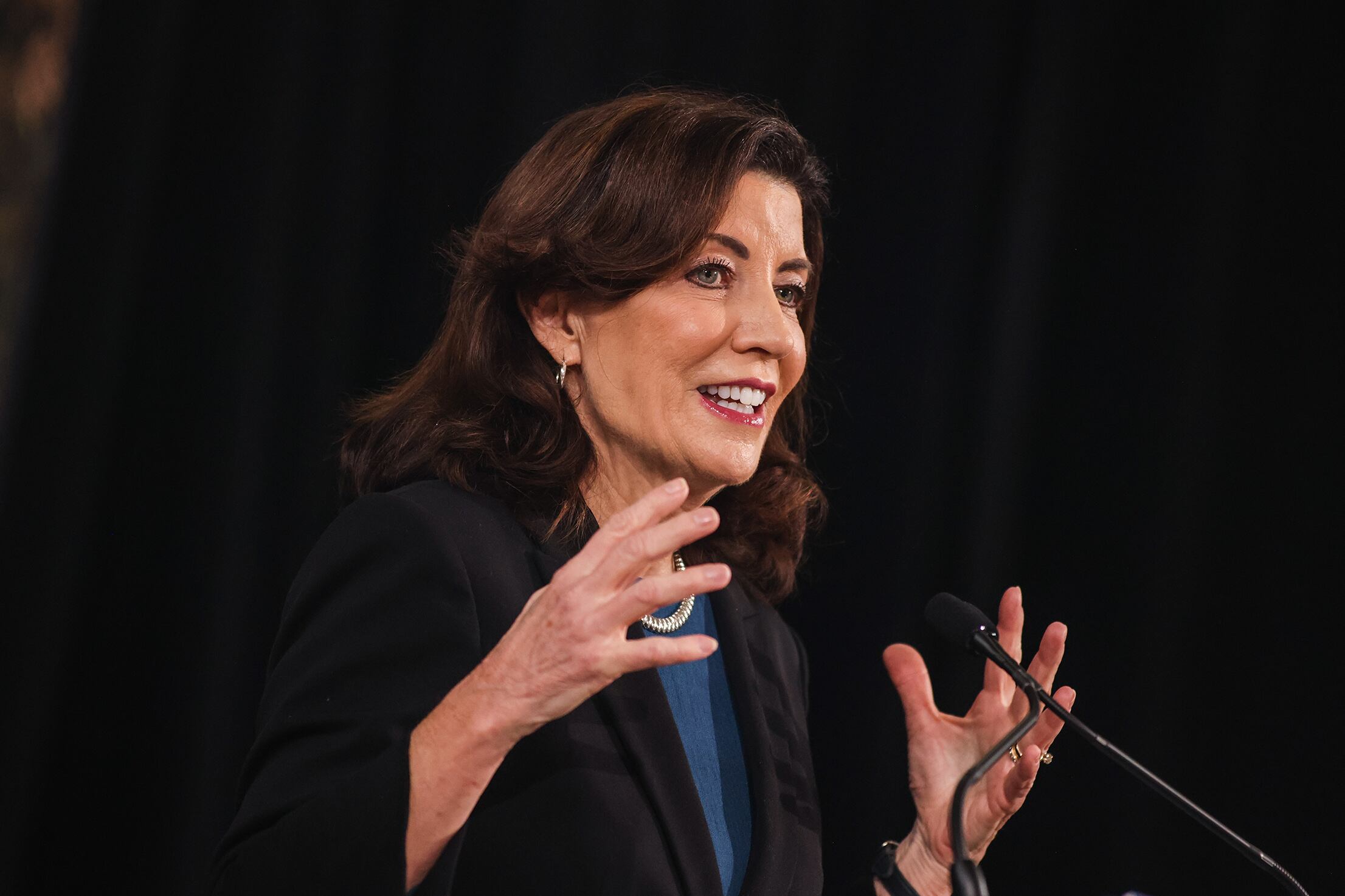 A woman with medium length brown hair holds her hands up while she speaks at a podium with a dark background.