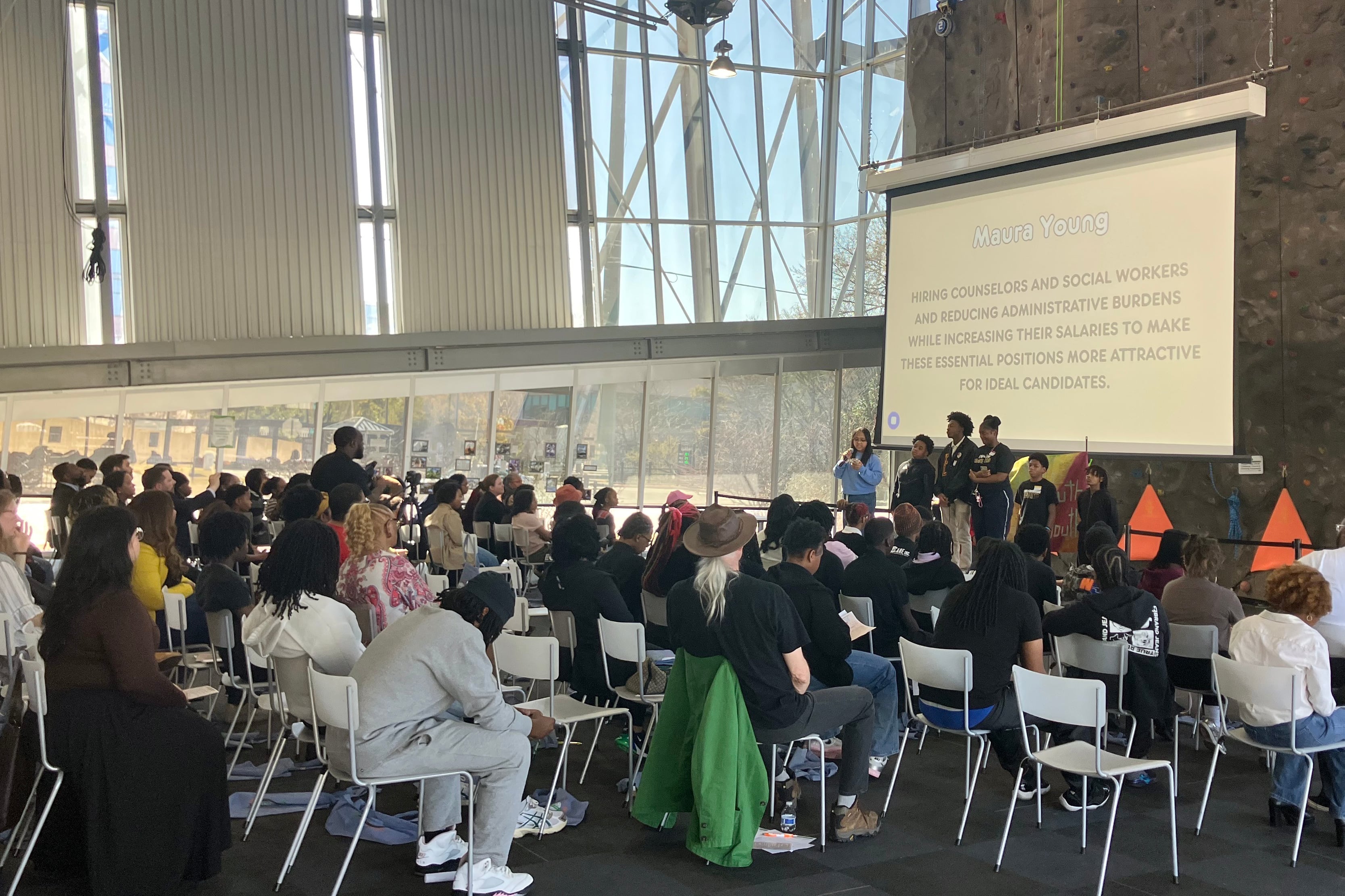 A photograph of a group of people sitting in chairs listening to a person speak all next to a climbing wall.