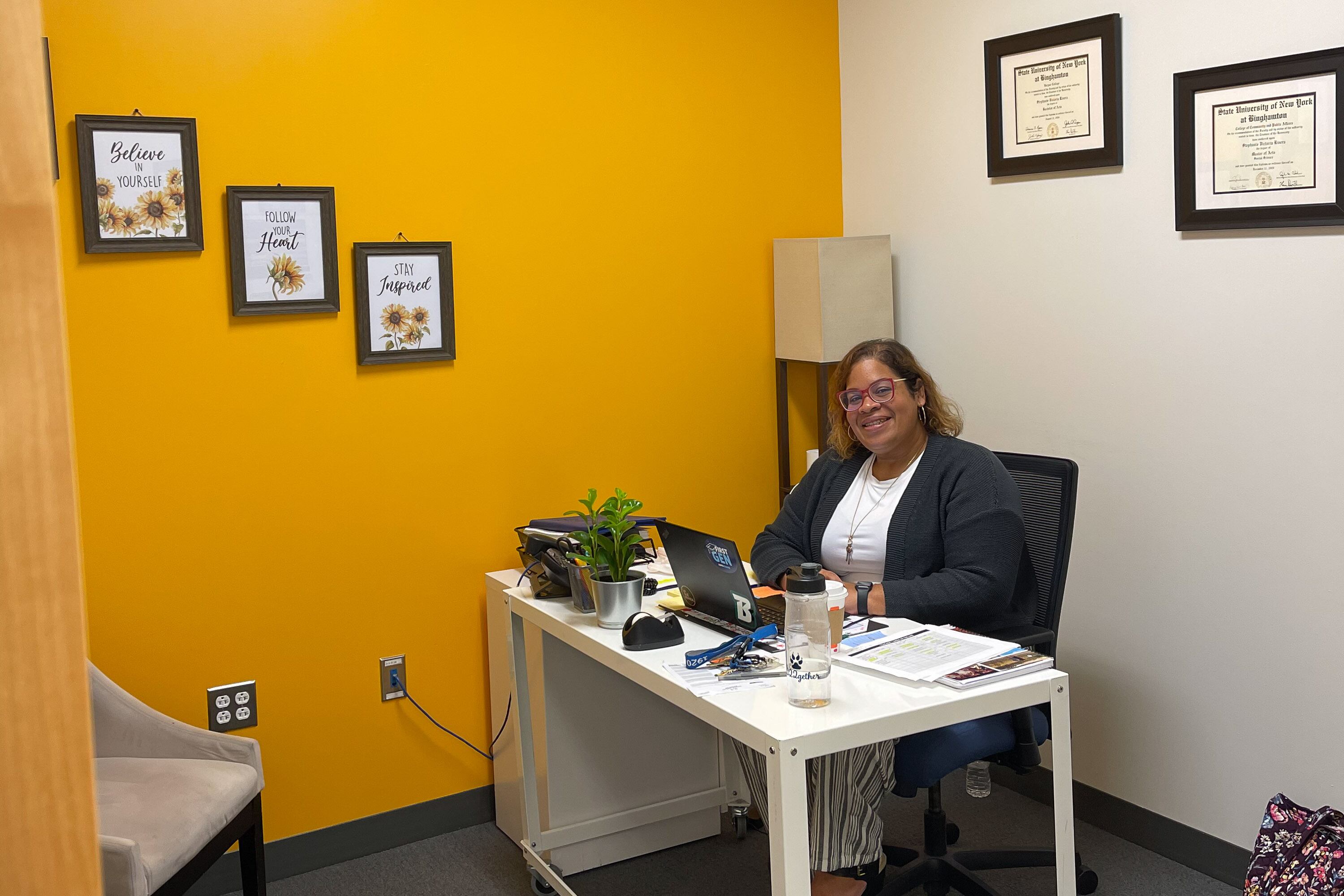 A woman wearing a black sweater and white shirt poses for a portrait at her desk. The room has one golden-colored wall and one white one, with framed pictures and diplomas on the walls.