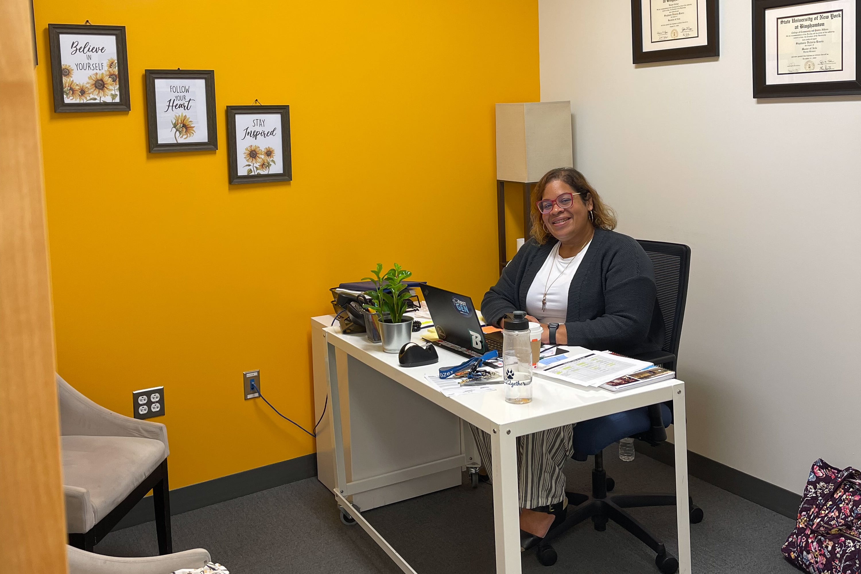 A woman wearing a black sweater and white shirt poses for a portrait at her desk. The room has one golden-colored wall and one white one, with framed pictures and diplomas on the walls.