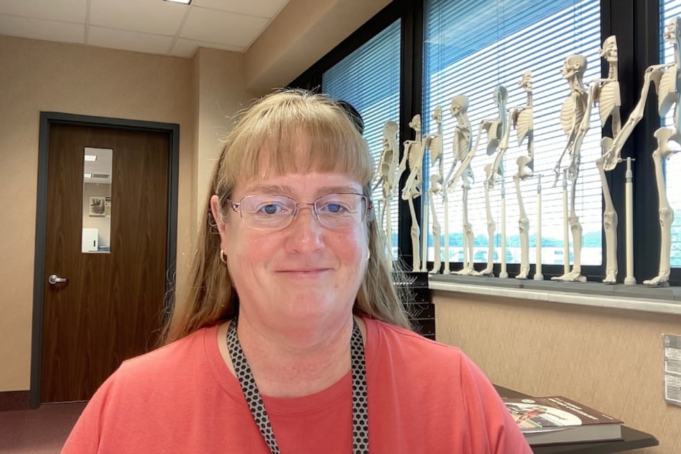 A photograph of a white woman wearing a salmon colored shirt next to a row of small, fake skeletons in an office.