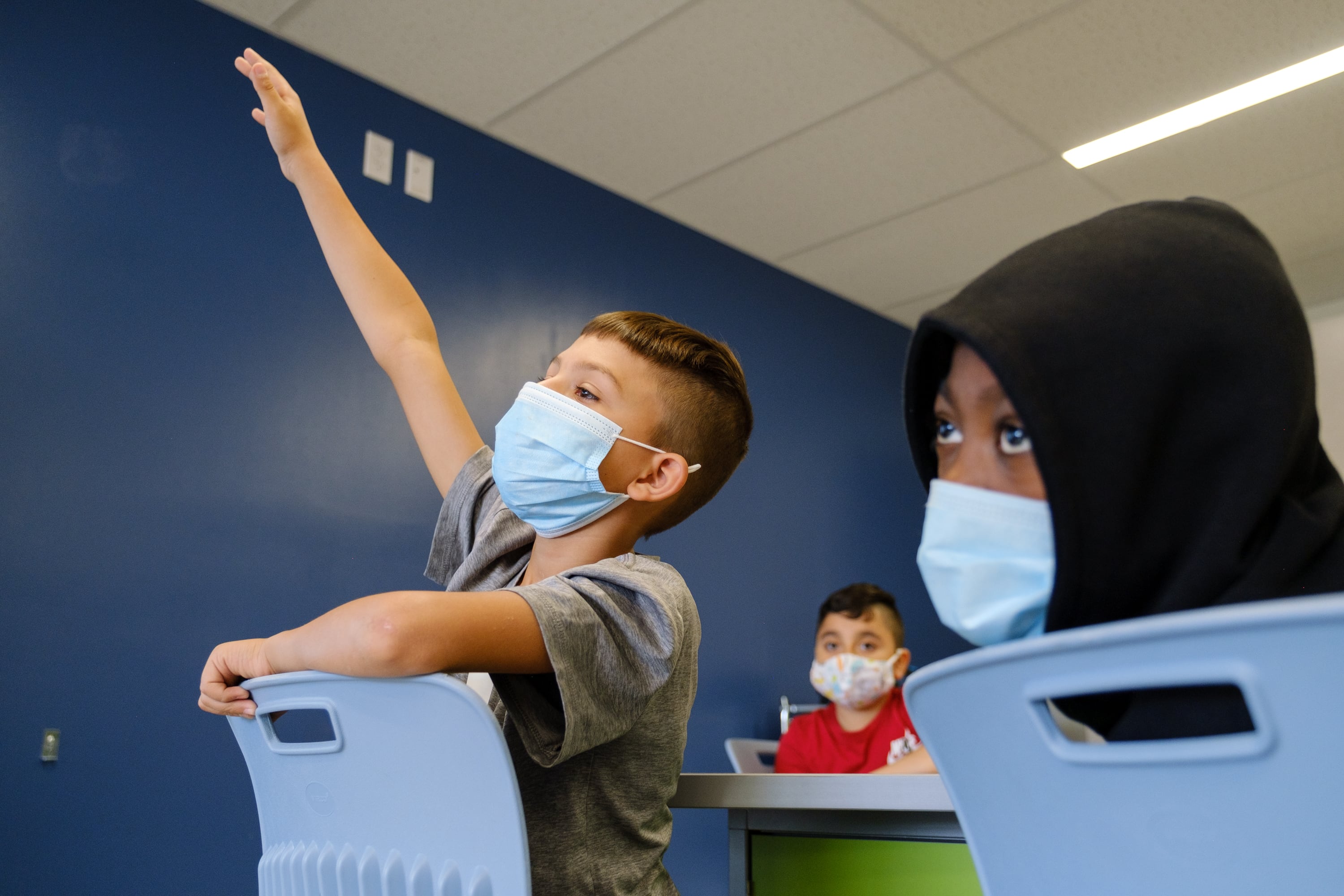 A young boy with short hair reaches his hand high in the air, straining to be called on in a classroom. Two other students look toward the front of the classroom as well. They are all wearing medical masks.