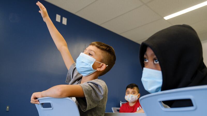 A young boy with short hair reaches his hand high in the air, straining to be called on in a classroom. Two other students look toward the front of the classroom as well. They are all wearing medical masks.