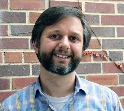Headshot of a man standing by a brick wall. He has a beard and is wearing a multicolored plaid shirt.