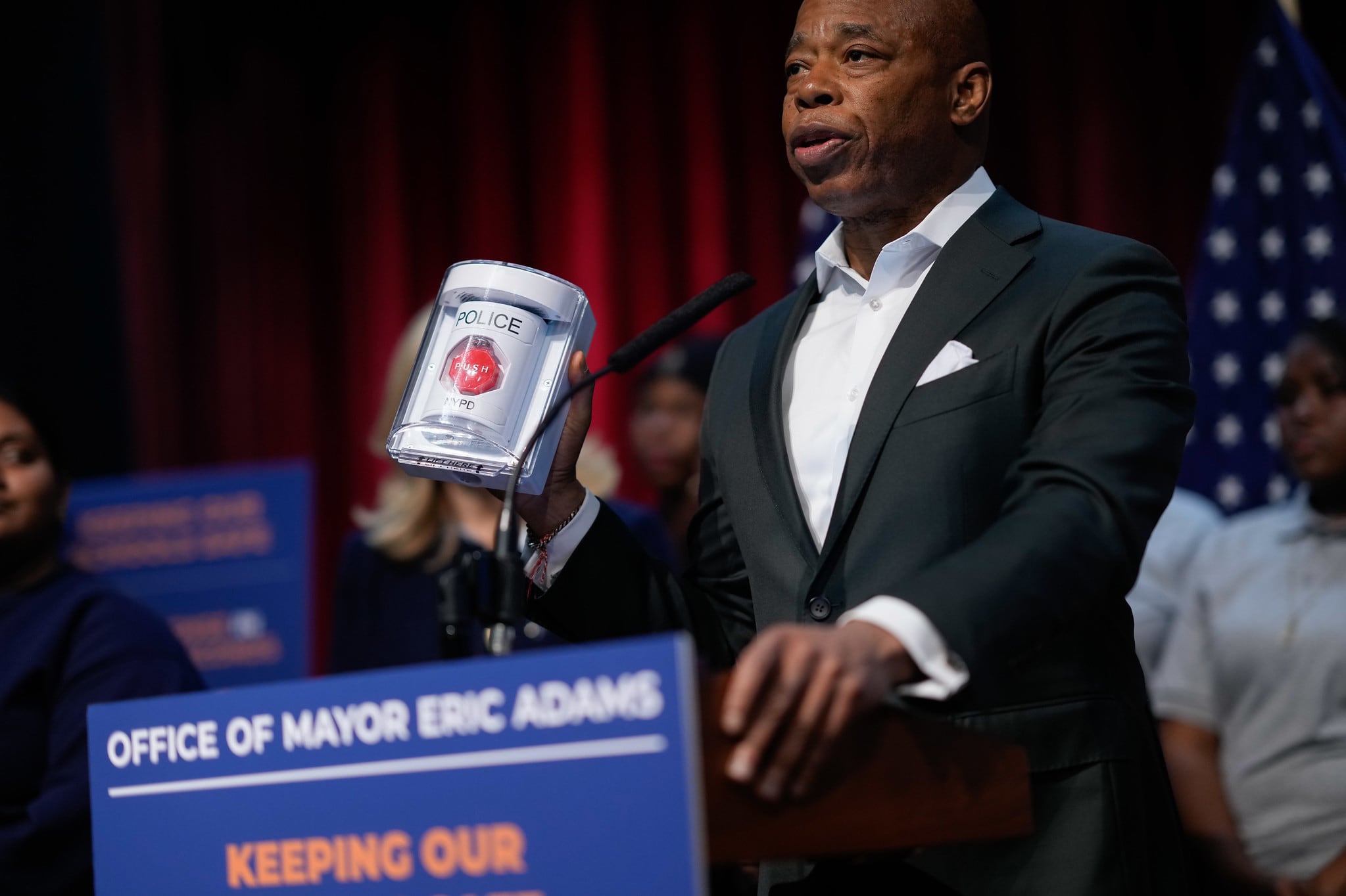 A photograph of a Black man in a suit holding up a device while speaking into a microphone from on stage with people in the background.