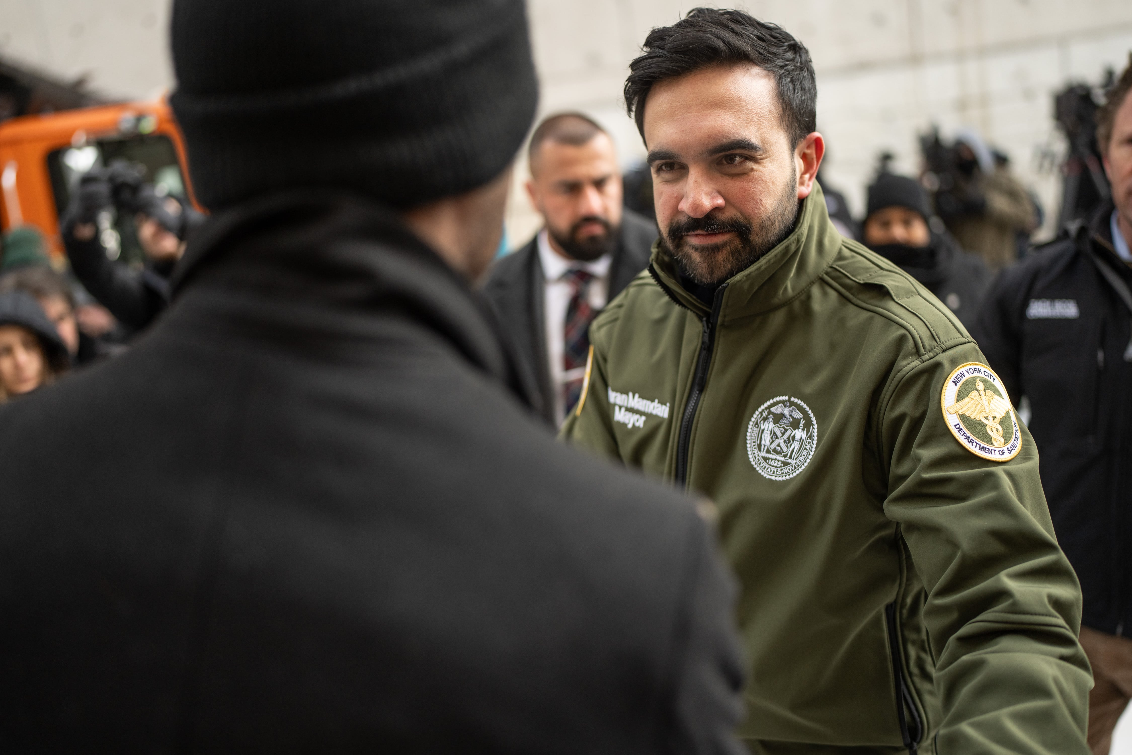 A man in an olive green coat greets another man.