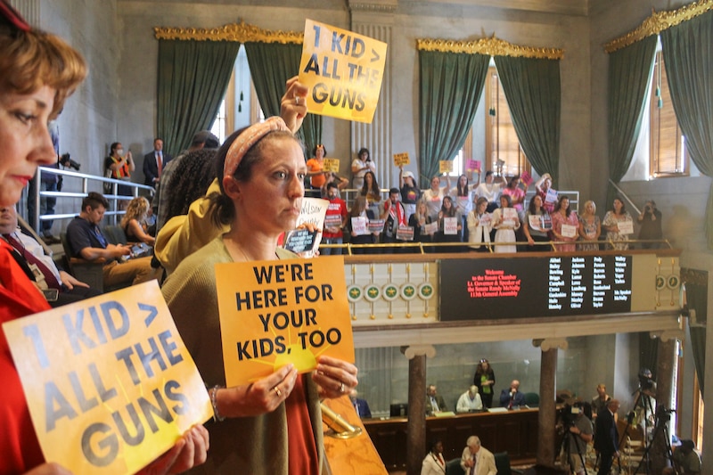 Women hold up signs of protest in a legislative chamber filled with people.
