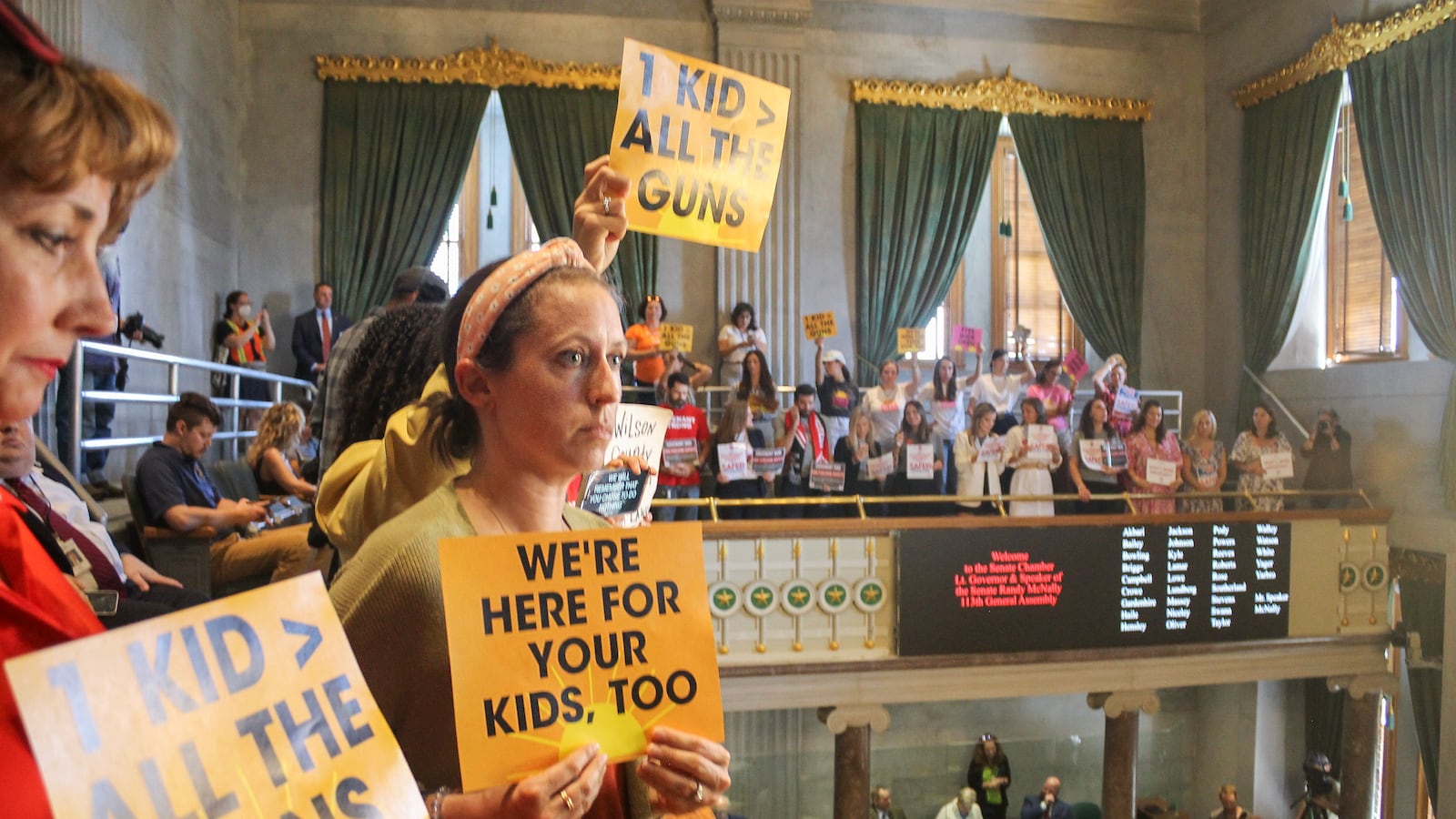 Women hold up signs of protest in a legislative chamber filled with people.