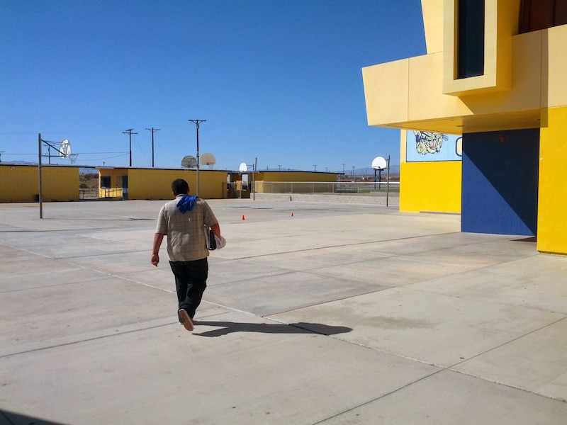 A student wearing dark pants and a tan shirt walks through an empty school courtyard with buildings and basketball courts in the background.