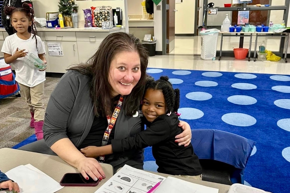 A woman in a gray jacket hugs a little girl at a table in front of a blue polka dotted carpet.