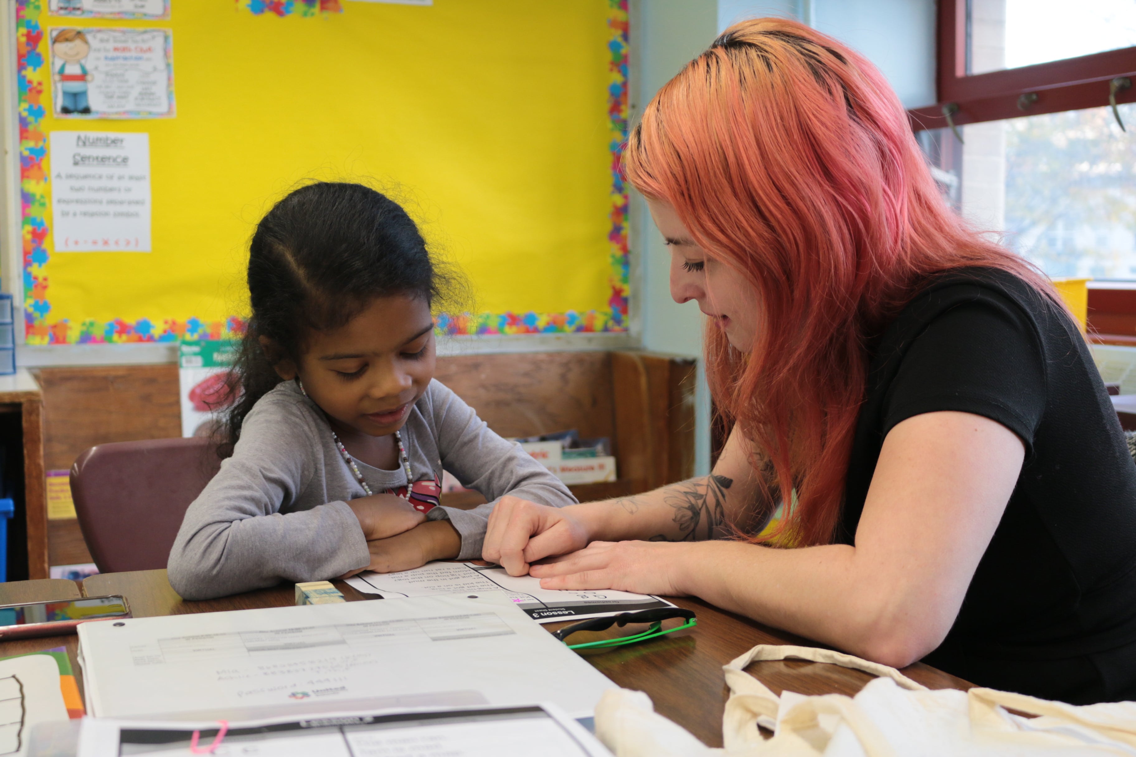 A young girl in a gray stop reads with a woman in a black top.