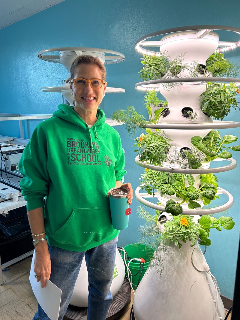A woman wearing a green hoodie stands next to a white hydroponic garden in a room with blue walls