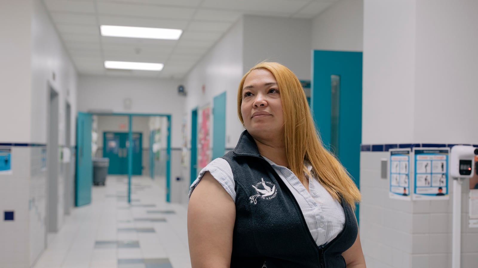 A woman with golden hair wearing a blue vest poses for a portrait in a school hallway.