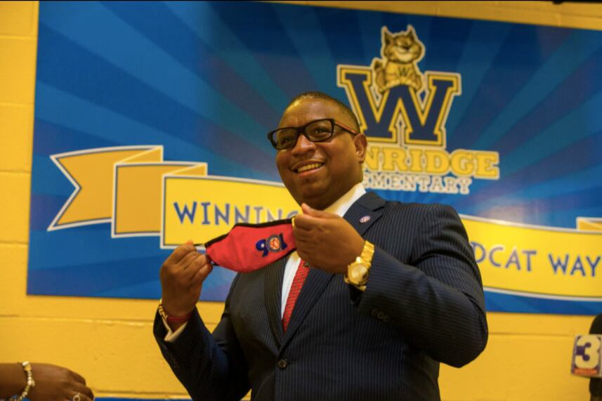 Superintendent Joris Ray stands in front of a school sign with a face mask in his hand with one of Shelby County Schools’ logos.